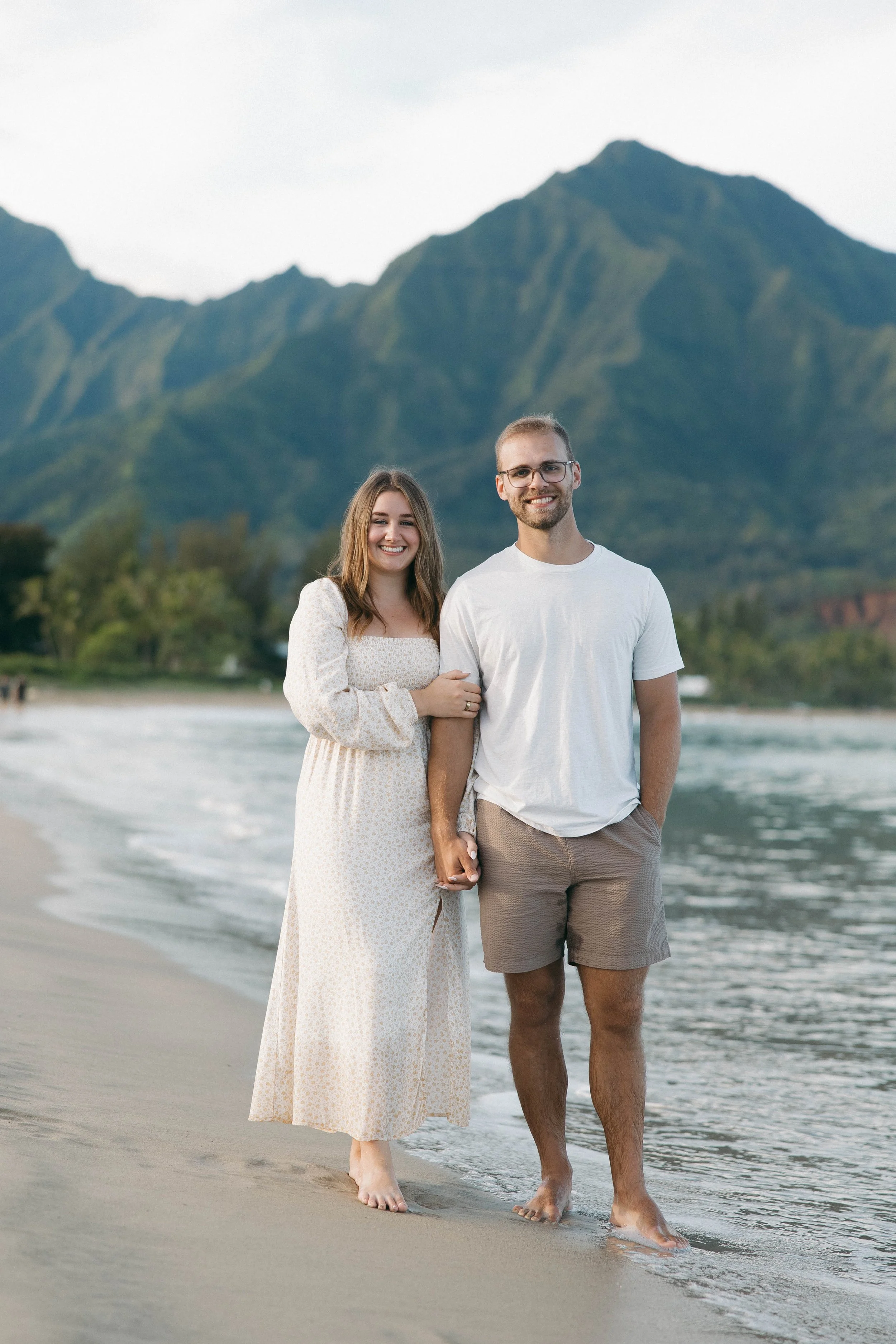 A smiling couple walking along the beach holding hands, with mountains in the background.
