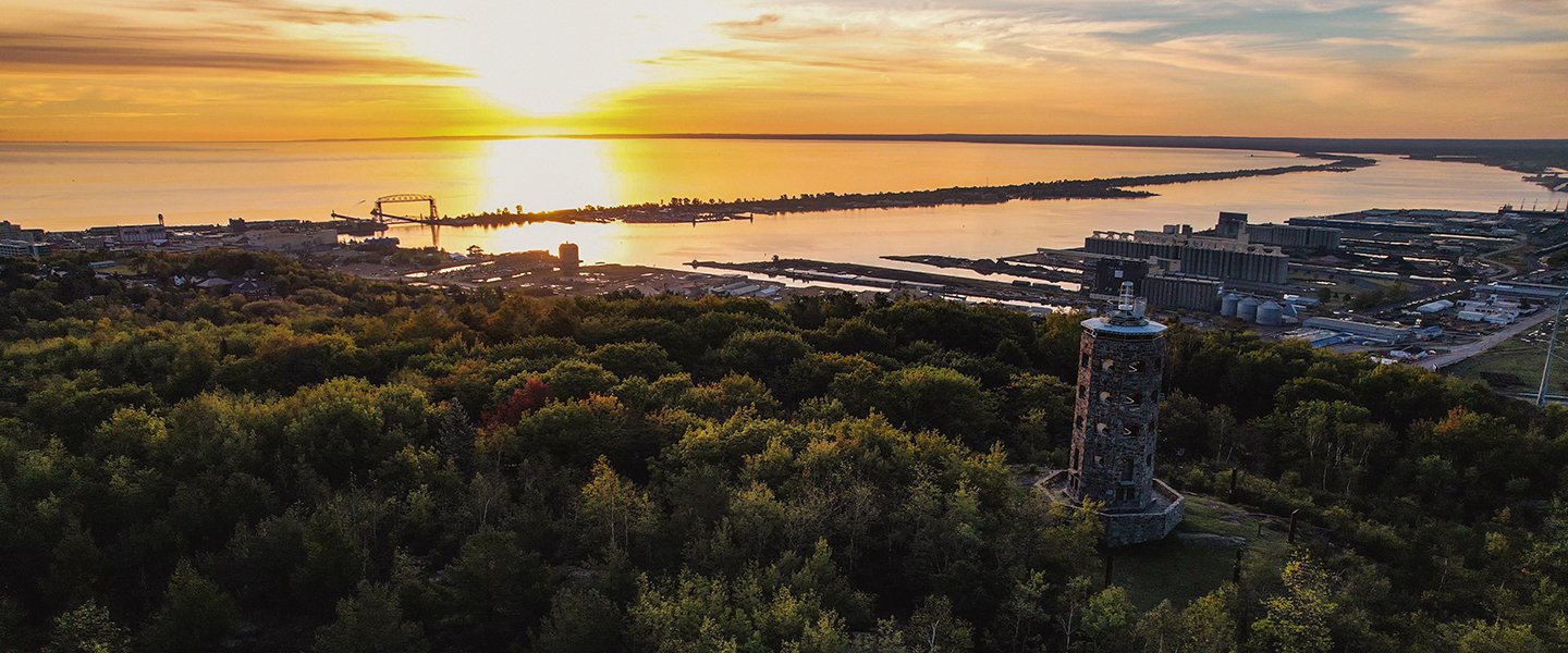 Sunset over a body of water with a lighthouse and a forested area in the foreground.