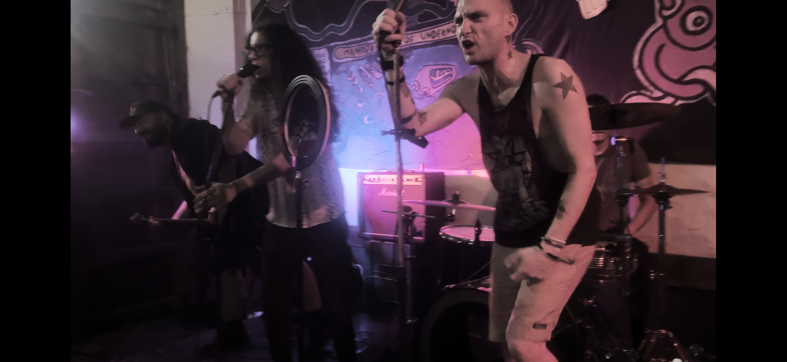 Three musicians perform on a dimly lit stage, one with a guitar, another singing into a microphone, and a drummer in the background, with pink and purple lighting.