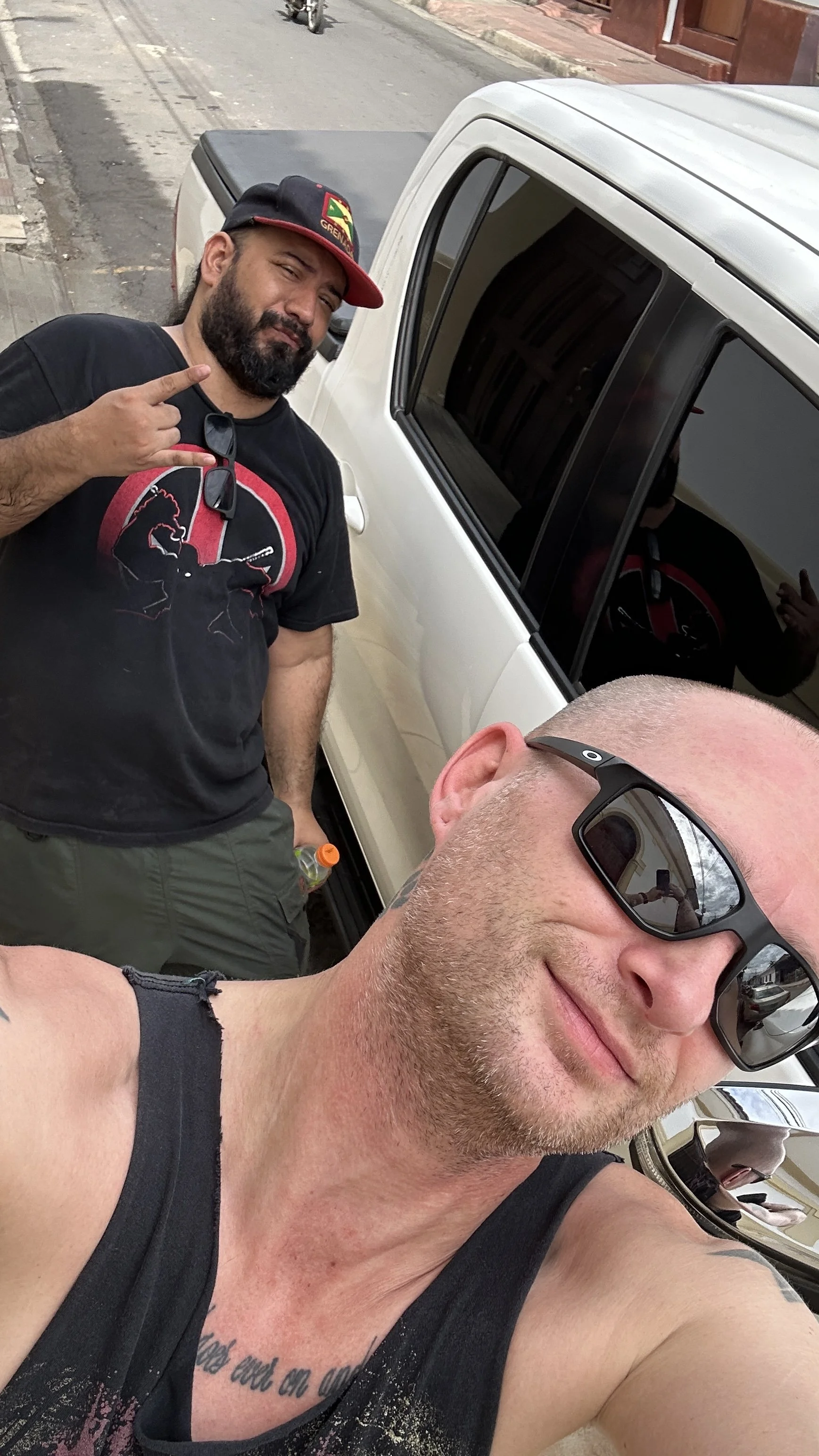 Two men taking a selfie next to a white pickup truck parked on the street. One man is in the foreground wearing sunglasses, smiling, and has a tattoo on his chest. The other man in the background is pointing towards the camera and wearing a black t-shirt with a graphic design and a cap.
