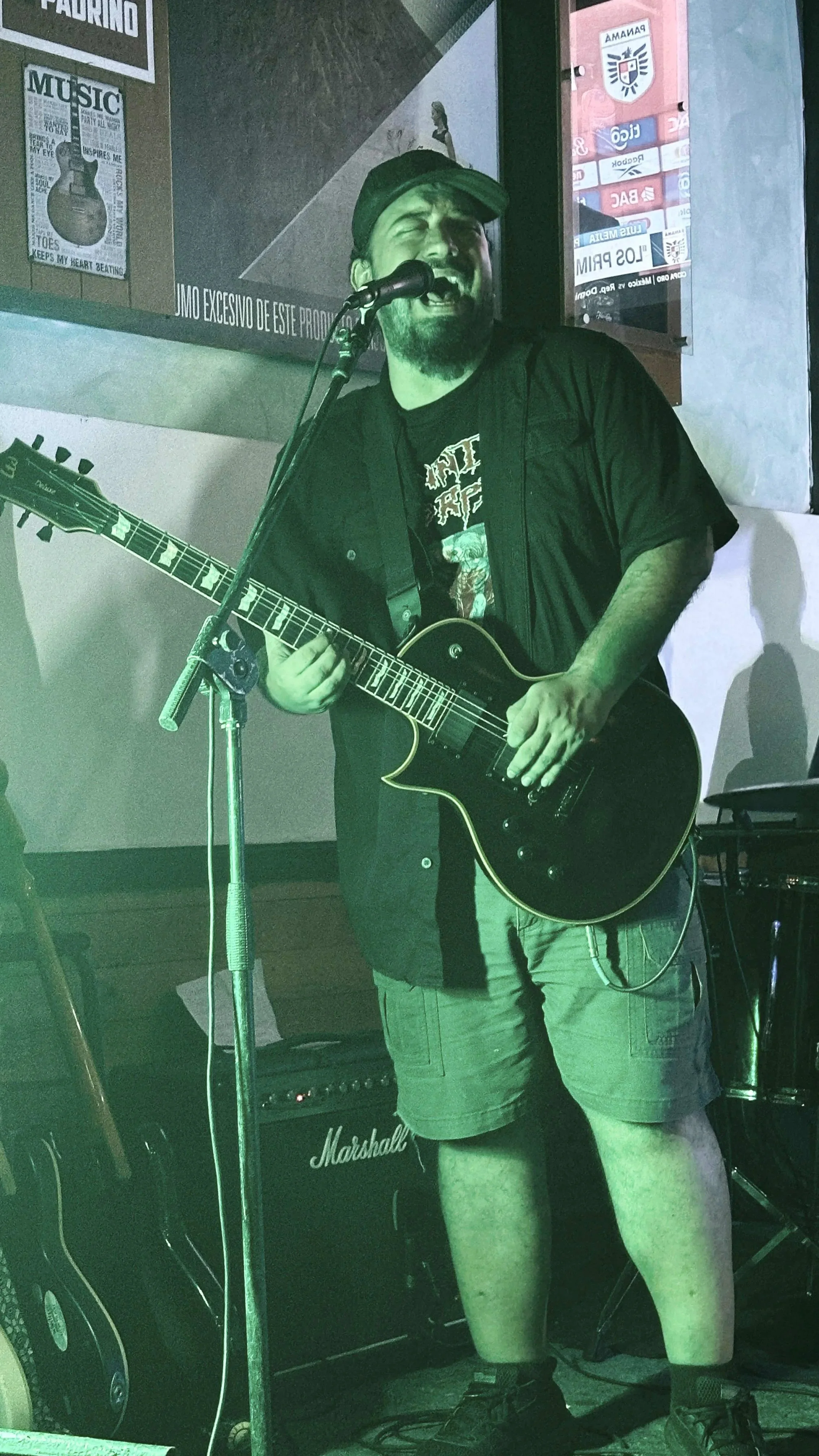 A man singing and playing an electric guitar in a music venue, with posters on the wall behind him.