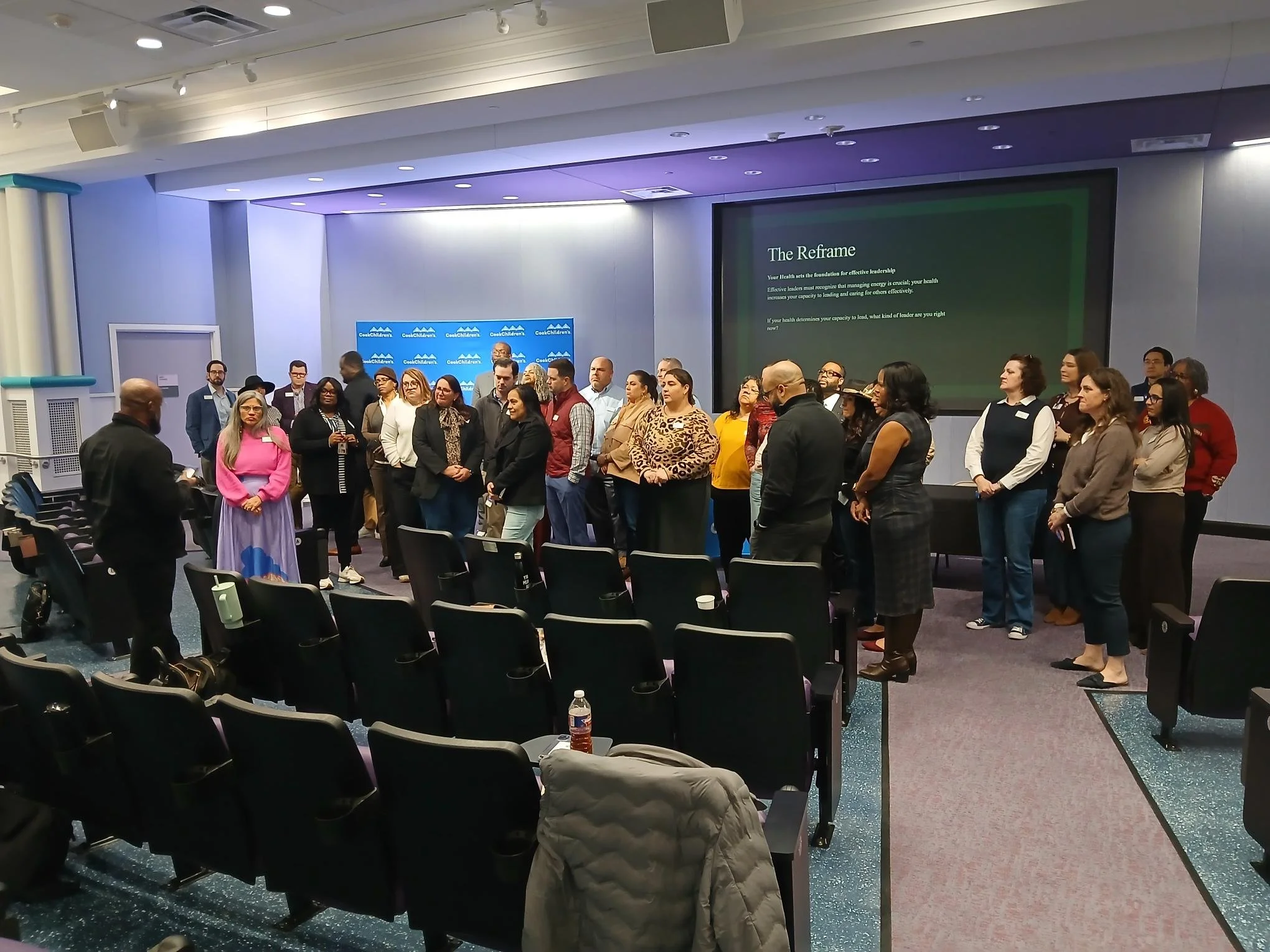 A group of people gathered in a conference room for a presentation or event, with a large screen displaying a slide titled 'The Reframe' and a blue backdrop with the logo 'Cool Children's'.