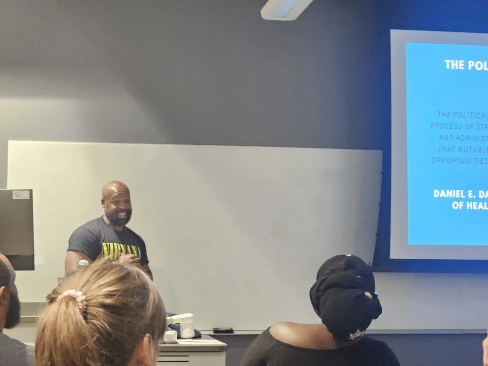 A man stands at the front of a classroom, giving a presentation to a group of students. The classroom has a projector screen displaying a slide, and there are students seated and listening.