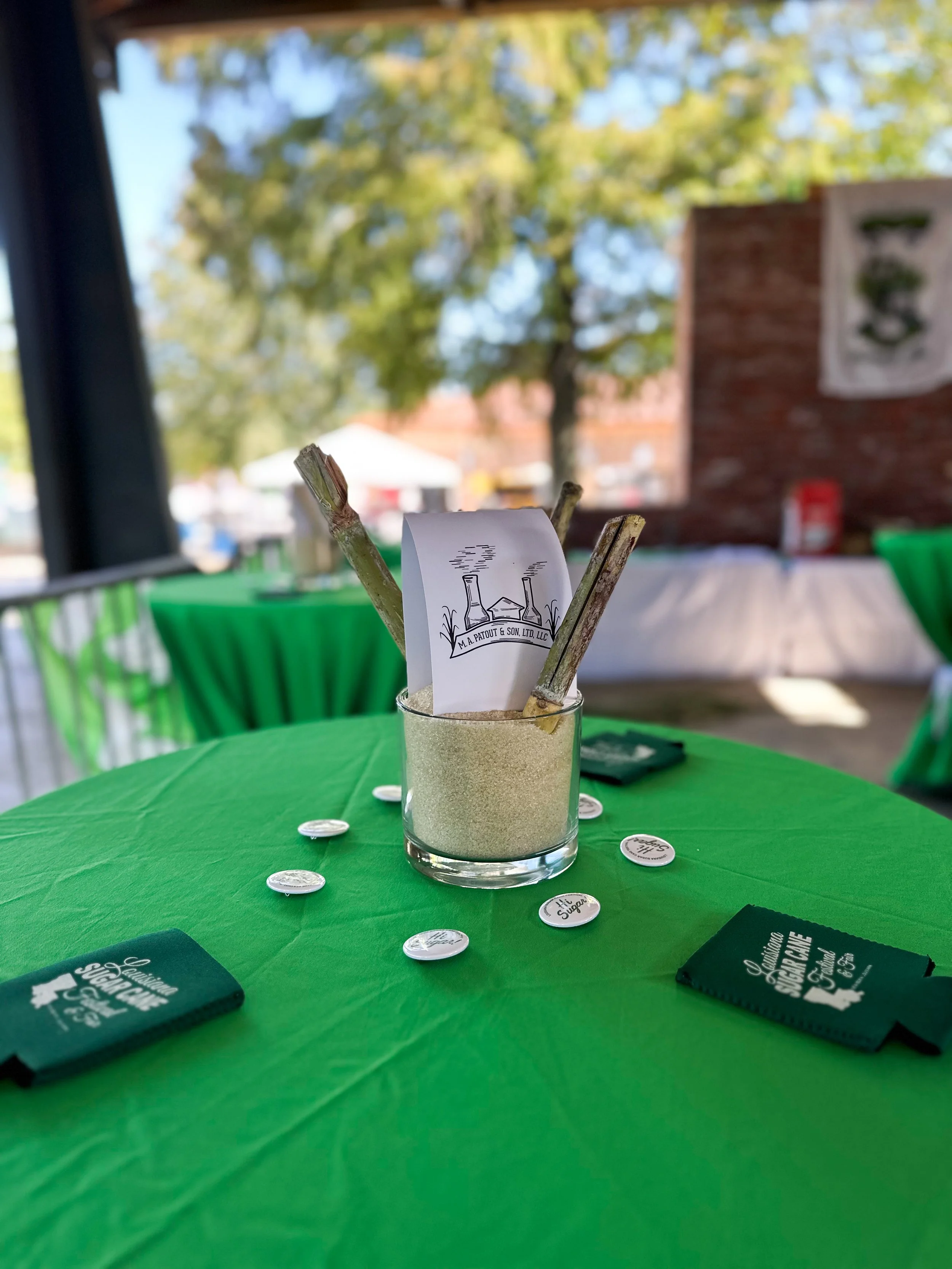 A glass filled with sand and two small wooden sticks holding a piece of paper with a logo. The table is covered with a green tablecloth and surrounded by circular tokens and black napkins with white text.