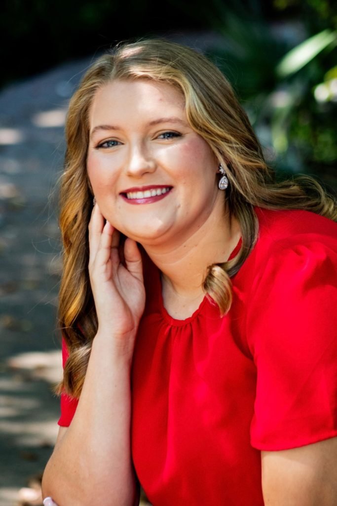 A woman with long blonde hair wearing a red top and earrings, smiling and resting her face on her hand outdoors.