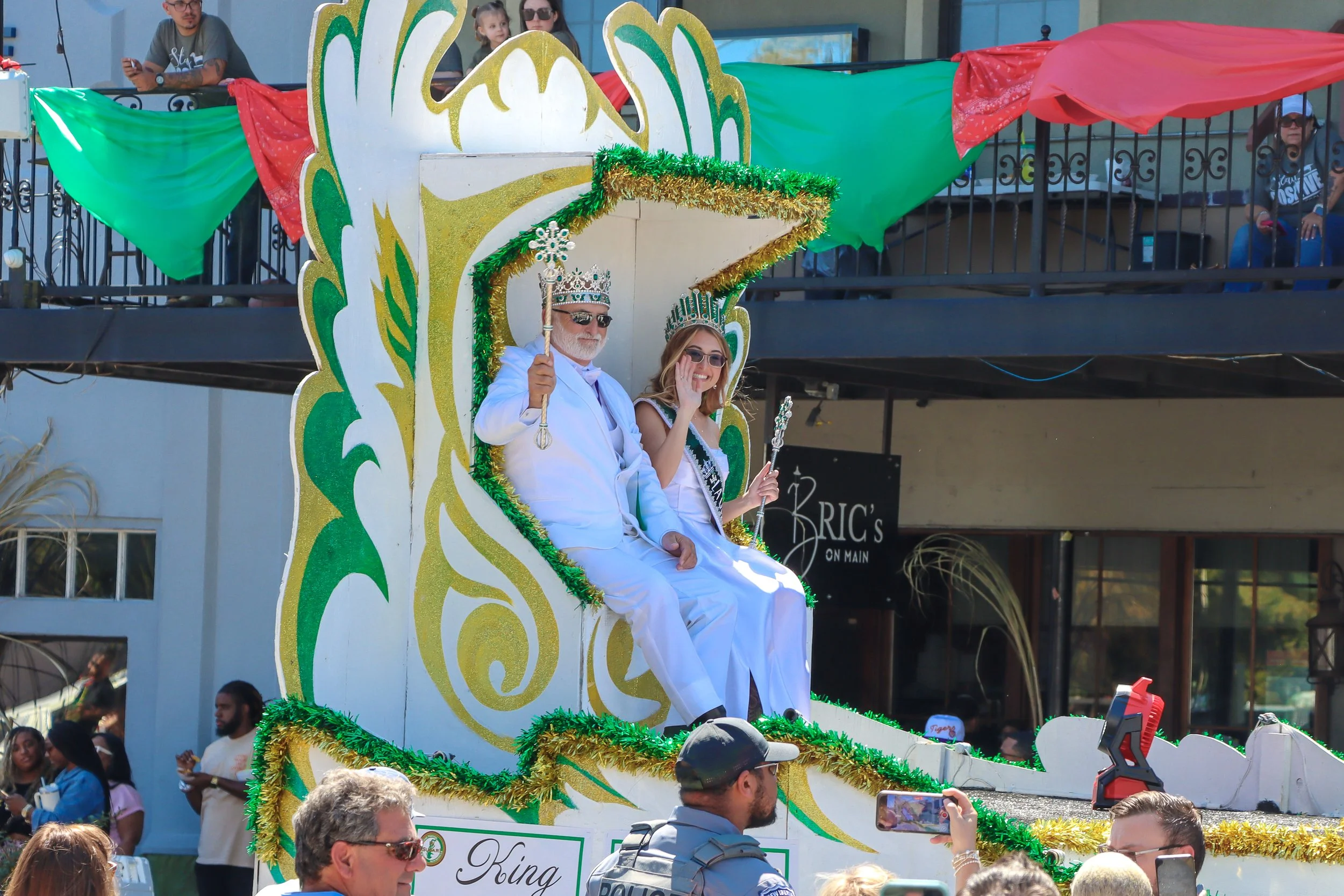 A decorated parade float with two individuals dressed as royalty sitting on it. The float is adorned with green, gold, and red decorations, including two crowns worn by the individuals. Spectators are gathered around, some taking photos, and others standing or talking along the street.