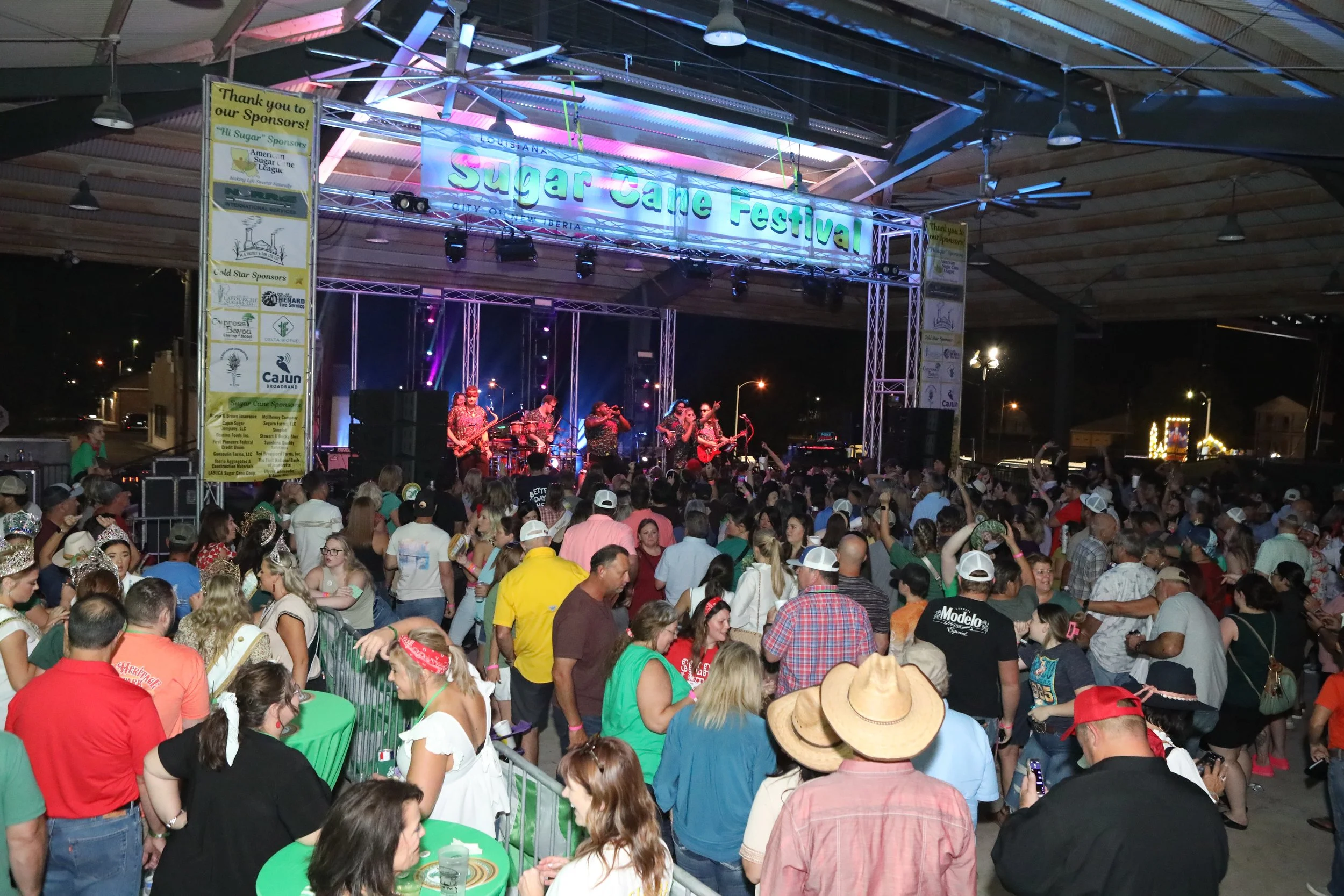 A crowded indoor event with a stage and live band performing. Attendees are standing and sitting, some wearing hats and casual clothes. The stage has a large sign hanging above that reads 'Sugar Cane Festival,' and there are banners on the sides thanking sponsors.