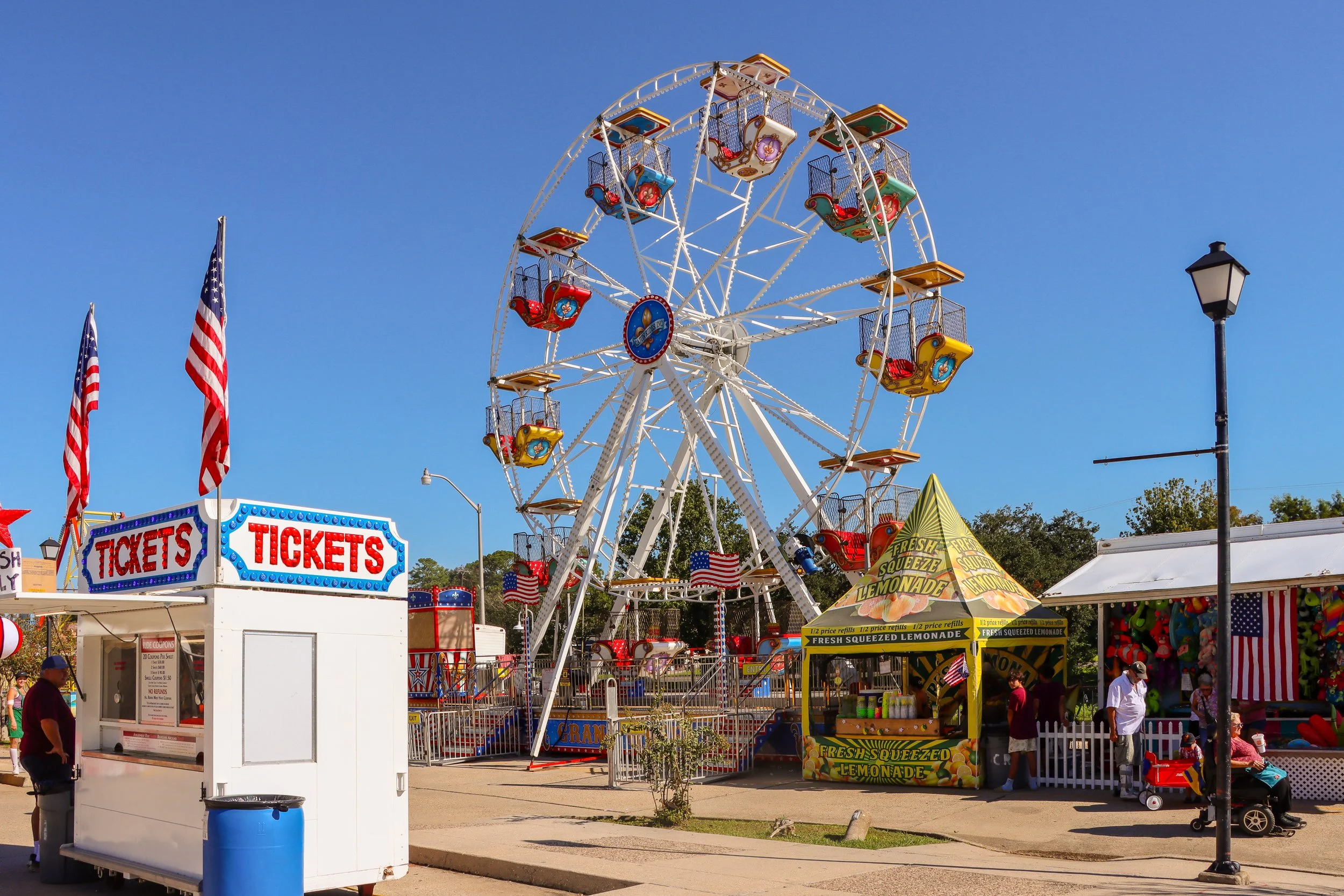 A colorful carnival with a Ferris wheel, ticket booth, and food stand selling lemonade, with American flags and people walking around on a sunny day.