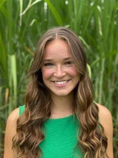 Young woman with long wavy brown hair, smiling, wearing a green sleeveless top outdoors with tall green grass in the background.