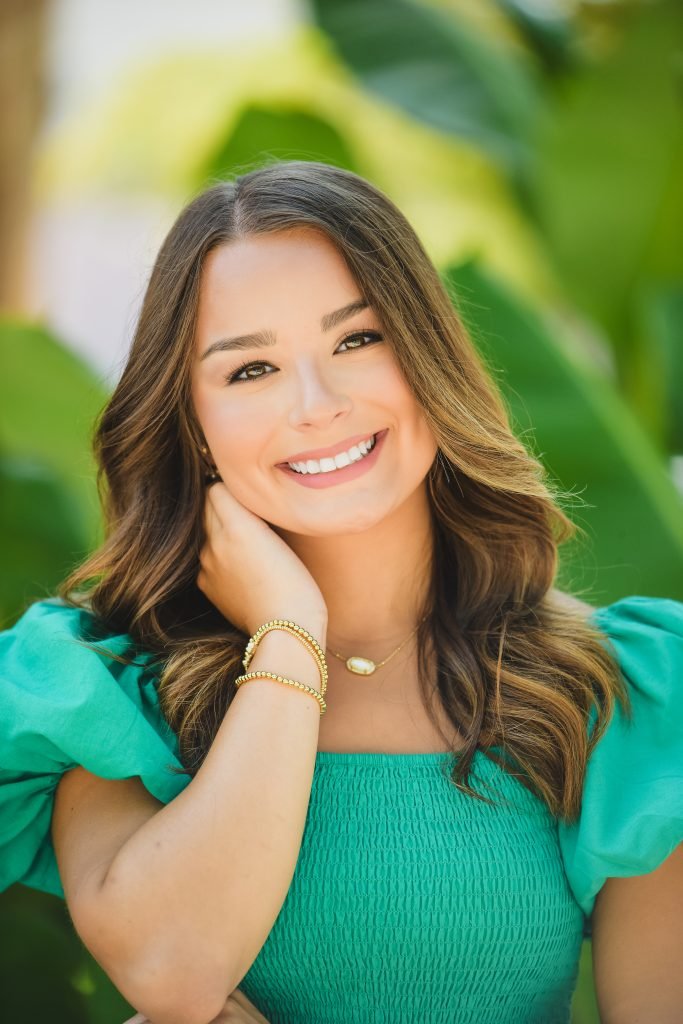A woman smiling outdoors, wearing a teal dress with puffed sleeves, gold jewelry, and long wavy hair.
