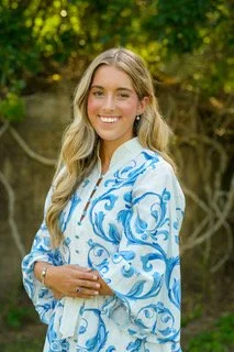 A young woman with long blonde hair smiling outdoors, wearing a white blouse with blue floral patterns, standing in front of green trees.