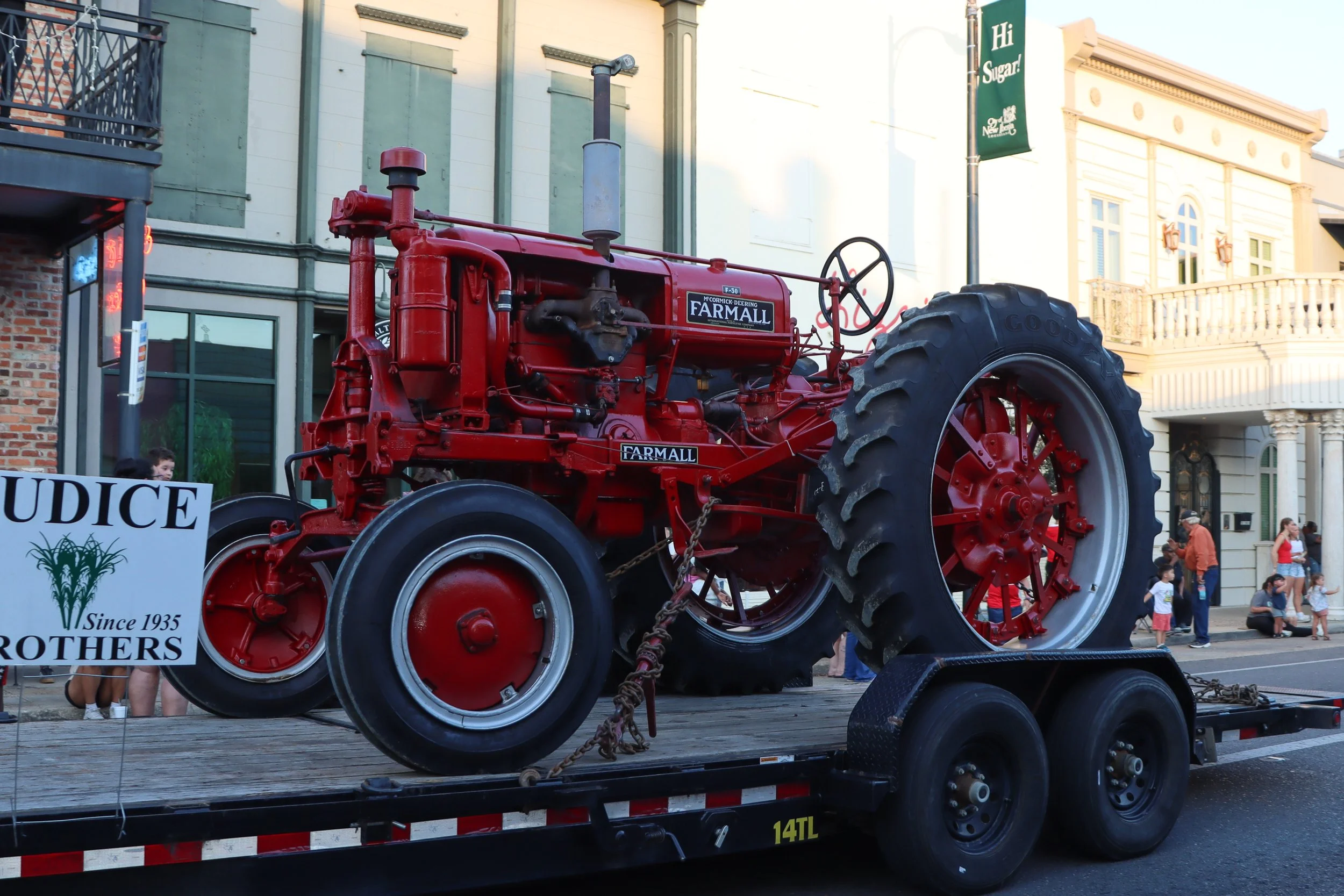 A large, vintage red tractor displayed on a flatbed truck during an outdoor event. There are people in the background, some children, and a building with light-colored architecture. A sign in the foreground reads "Since 1935" and includes a graphic of green wheat.