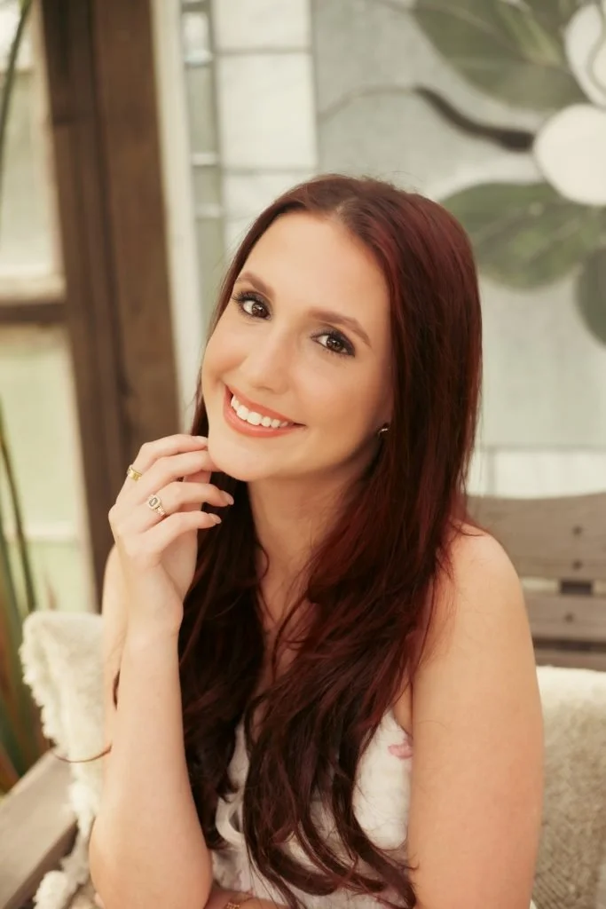 A woman with long red hair smiling while resting her chin on her hand in a cozy indoor setting.