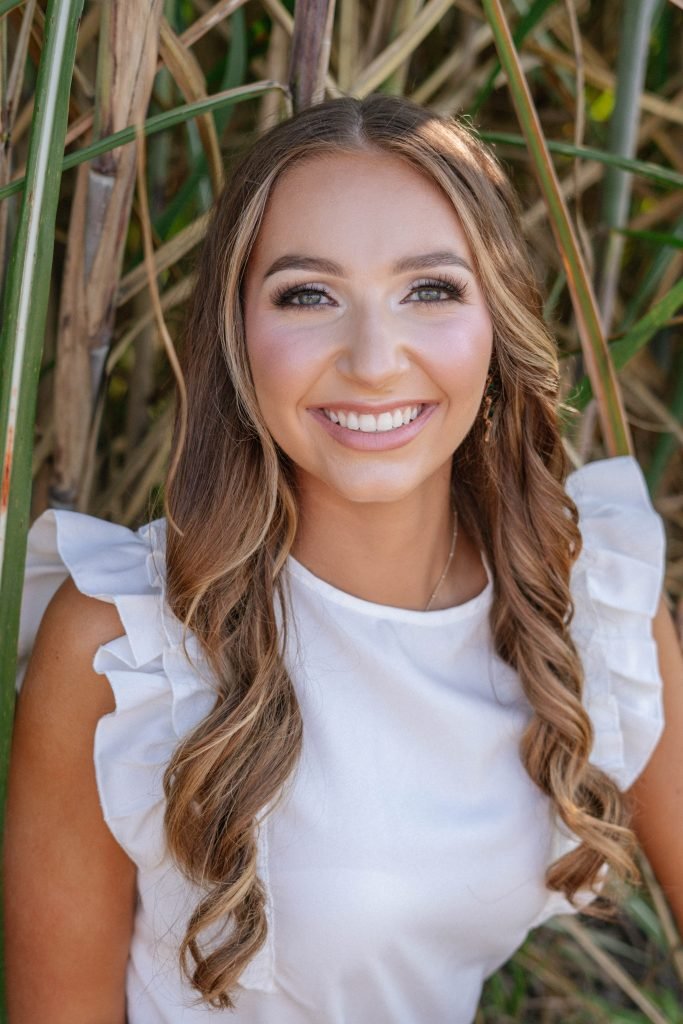 A young woman with long, wavy brown hair and light makeup smiling outdoors among tall grass or reeds, wearing a white top with ruffle sleeves.