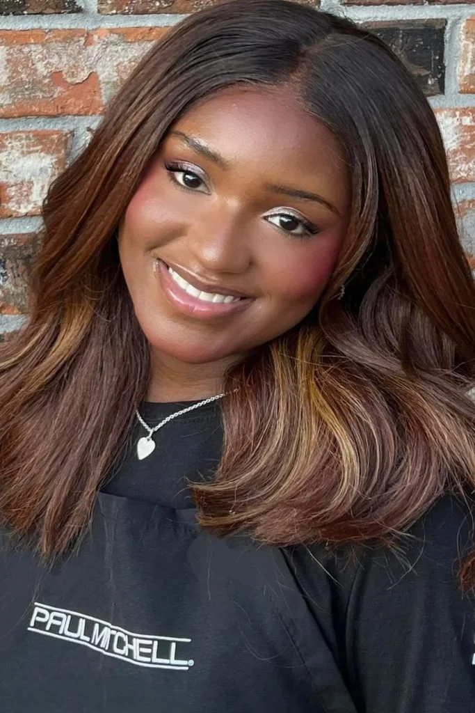 Smiling woman with long, layered brown hair wearing a black shirt with white text and a silver heart necklace, standing in front of a brick wall.