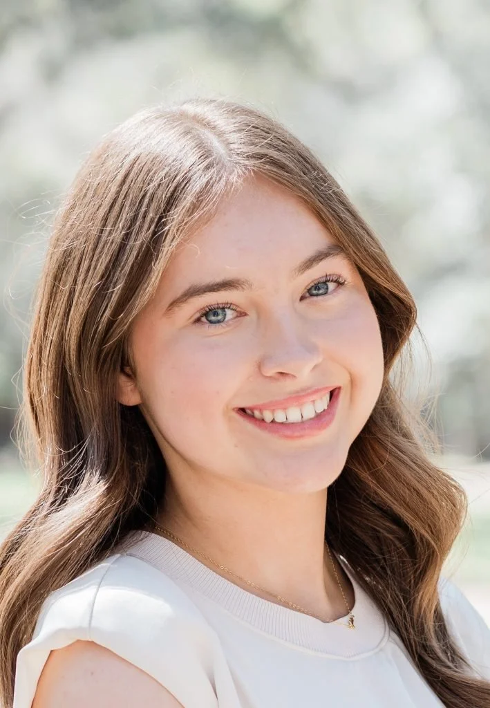 A young woman smiling outdoors with sunlight and blurred greenery in the background.