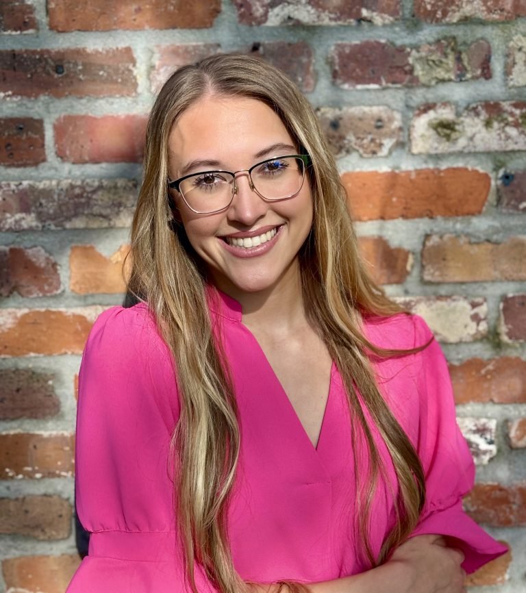 A young woman with long blonde hair and glasses, smiling in front of a rustic brick wall, wearing a bright pink blouse.