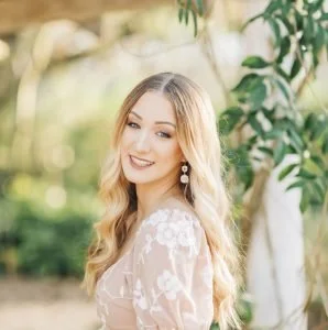 A young woman with long blonde hair smiling outdoors with greenery and trees in the background.