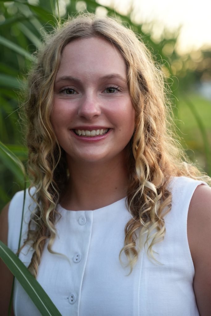 A smiling young woman with curly blonde hair, wearing a white sleeveless top, outdoors with greenery and sunlight in the background.
