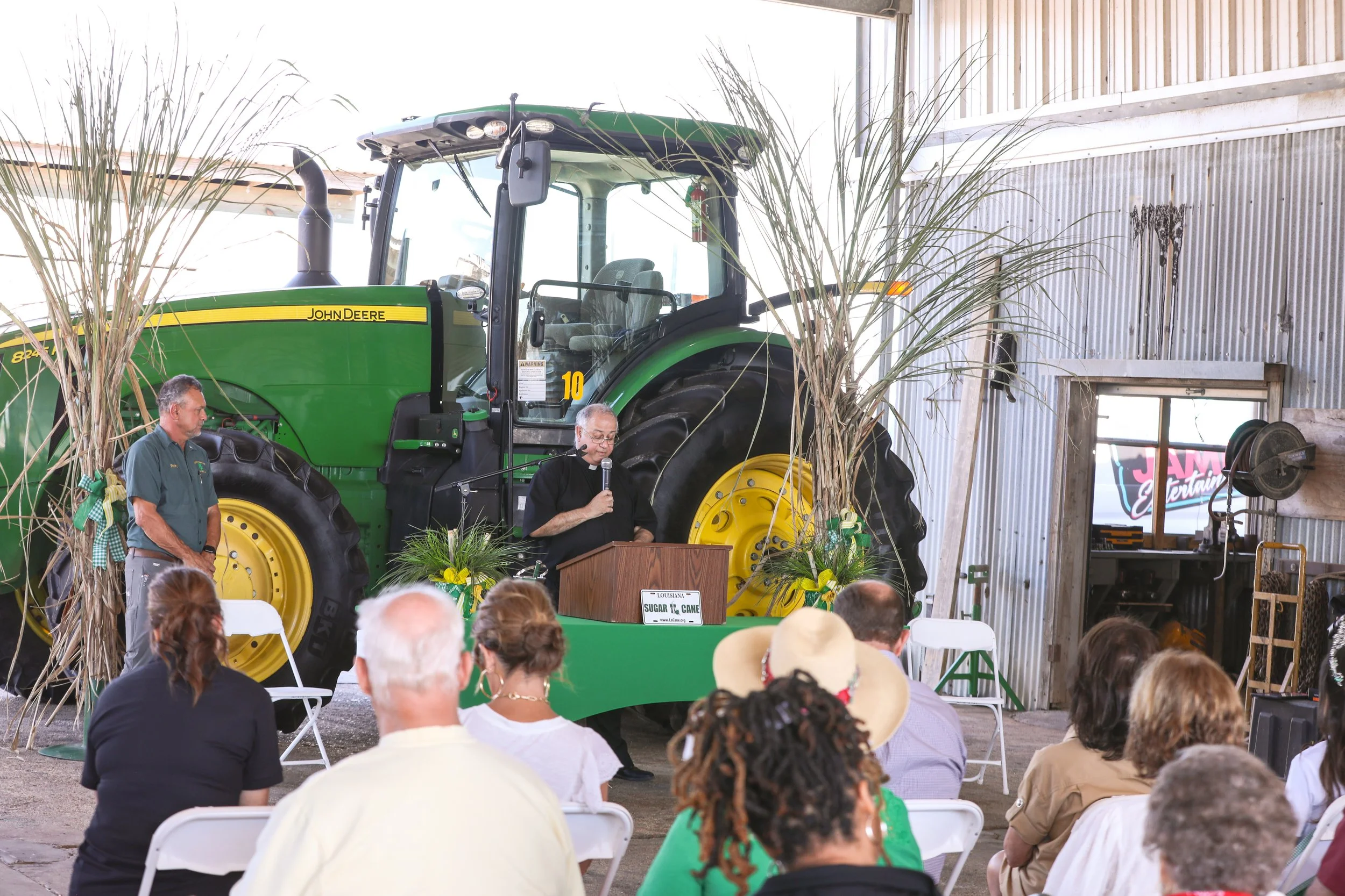 A man with gray hair speaking at a podium with a microphone, surrounded by an audience, with a green John Deere tractor in the background inside a metal building.