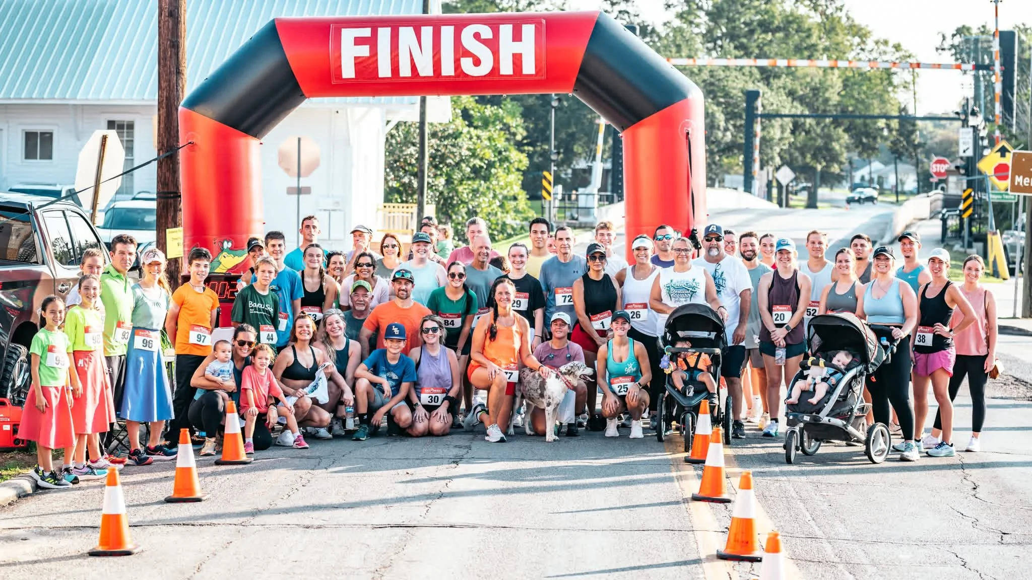 Group of people at the finish line of a race, standing under an archway with a red 'FINISH' sign, some with race bibs, in a sunny outdoor setting with trees and traffic signs nearby.