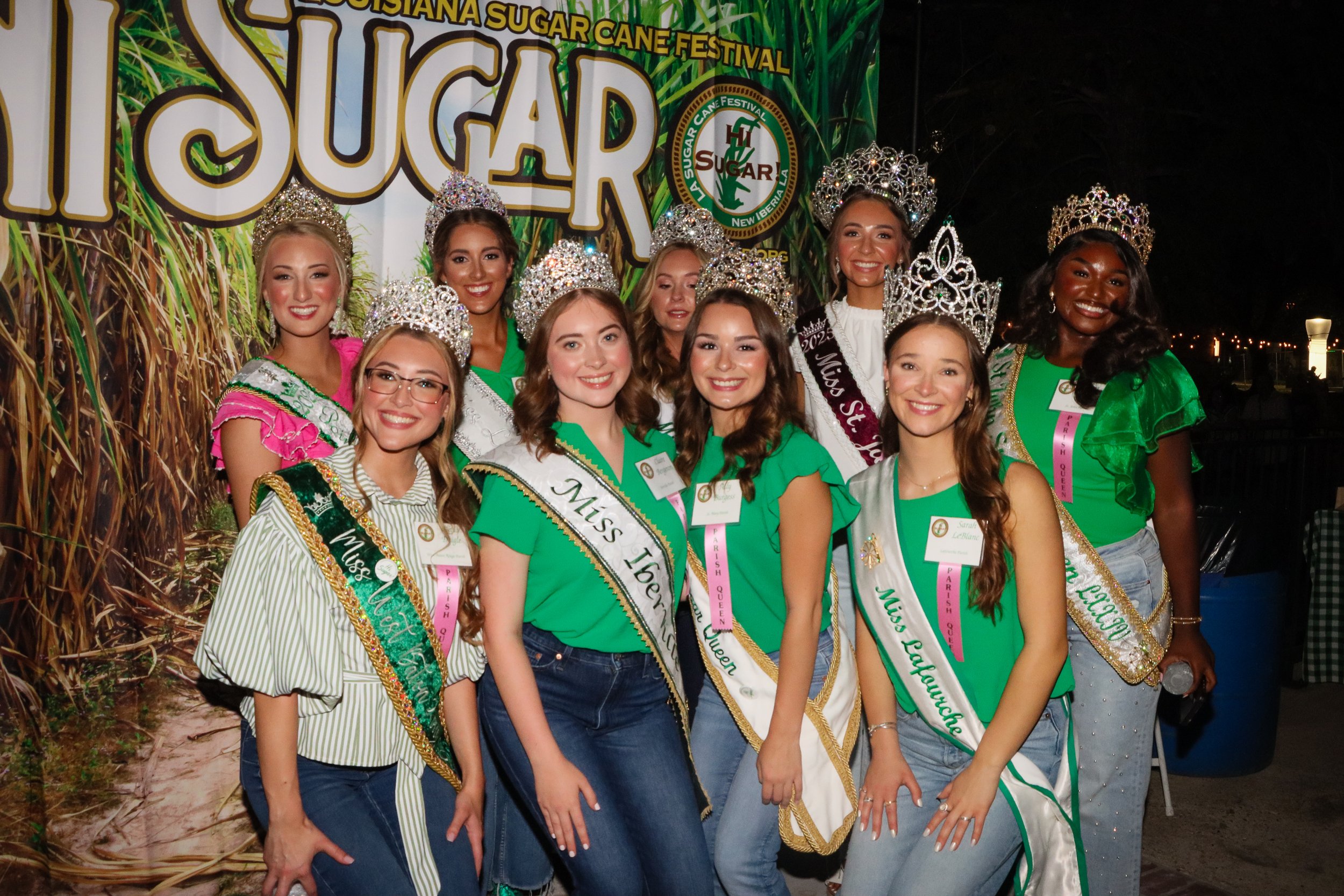 Group of young women dressed in colorful outfits and wearing crowns, standing in front of a large sign that reads 'Louisiana Sugar Cane Festival' with a sugarcane field in the background.