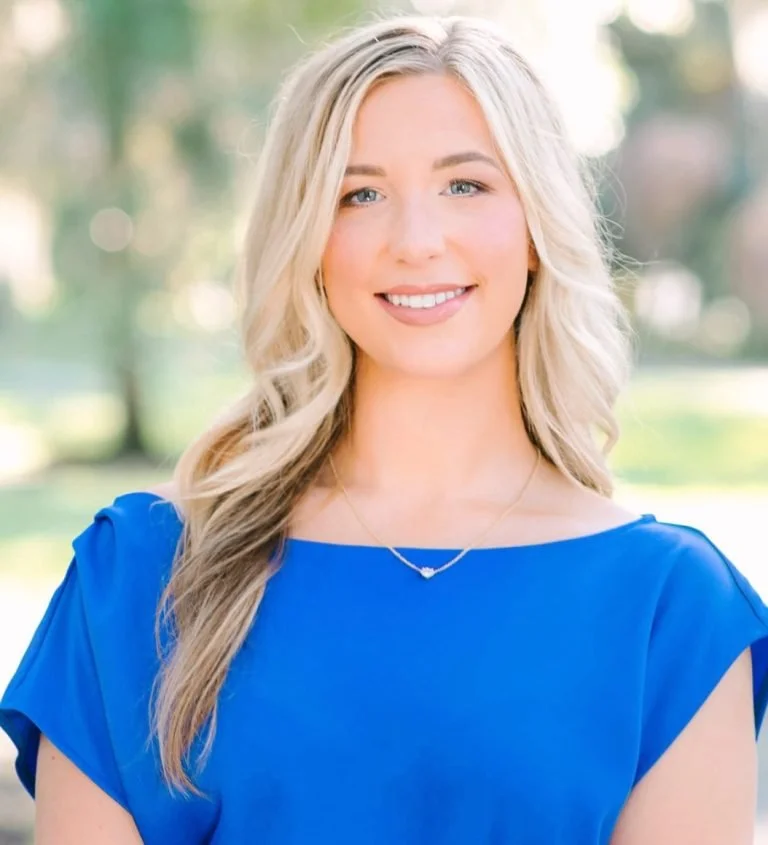 A young woman with long, wavy blonde hair and blue eyes smiling outdoors, wearing a blue top and a delicate necklace.
