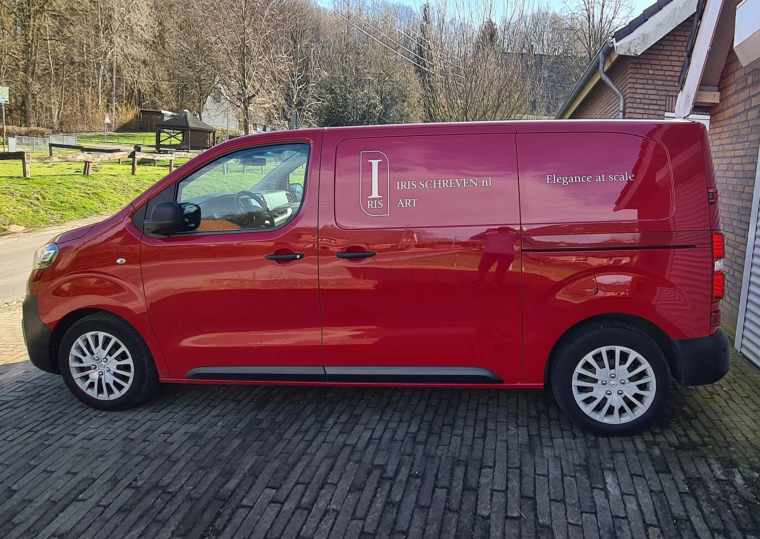 Red commercial van parked on cobblestone driveway next to brick building with garden and trees in the background.