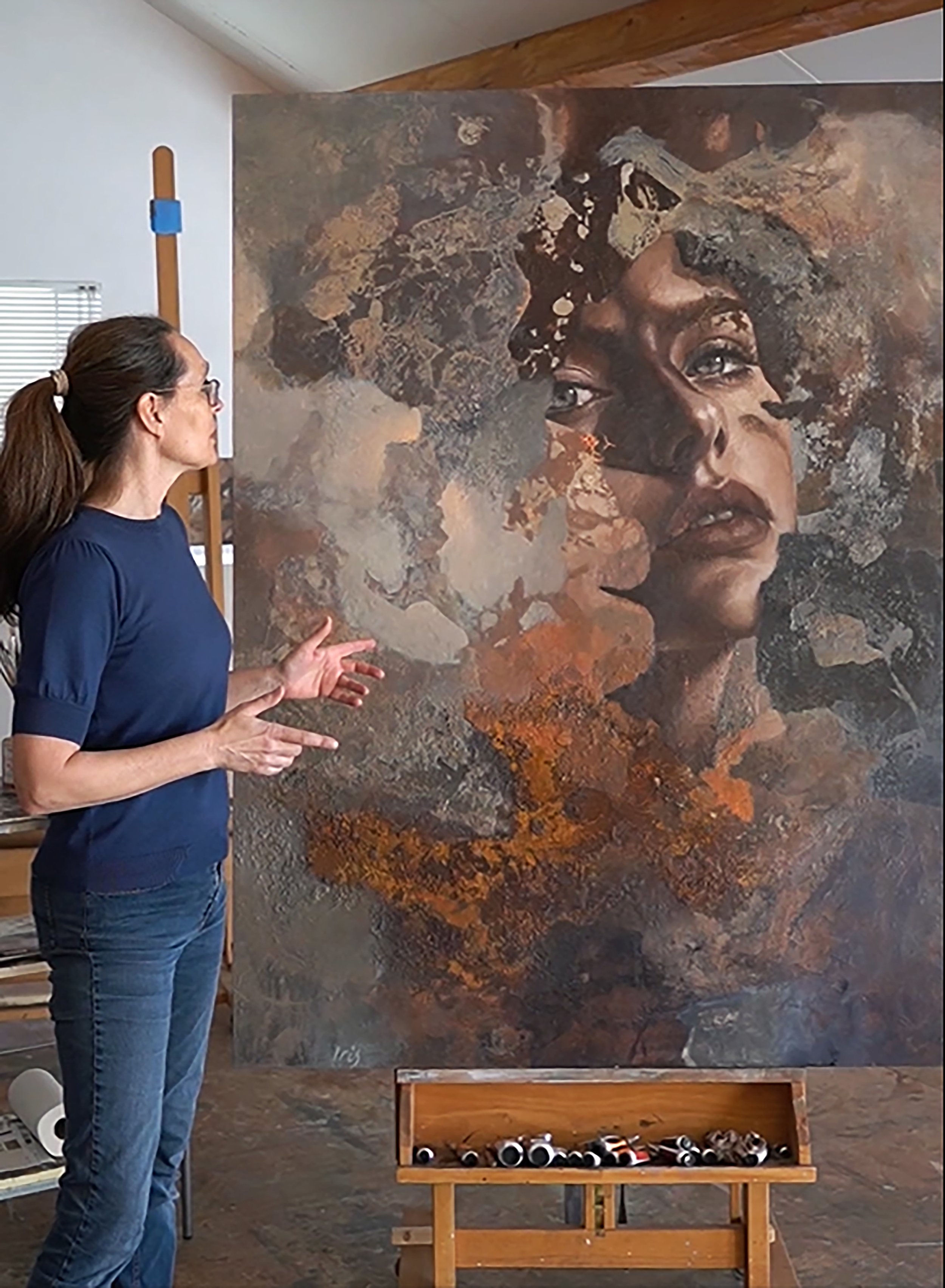 A woman with glasses and a ponytail stands in front of a large abstract artwork of a woman’s face with expressive eyes and open lips, in an art studio with a wooden floor and white walls. A wooden box with black and orange rolled papers is visible in front of her.