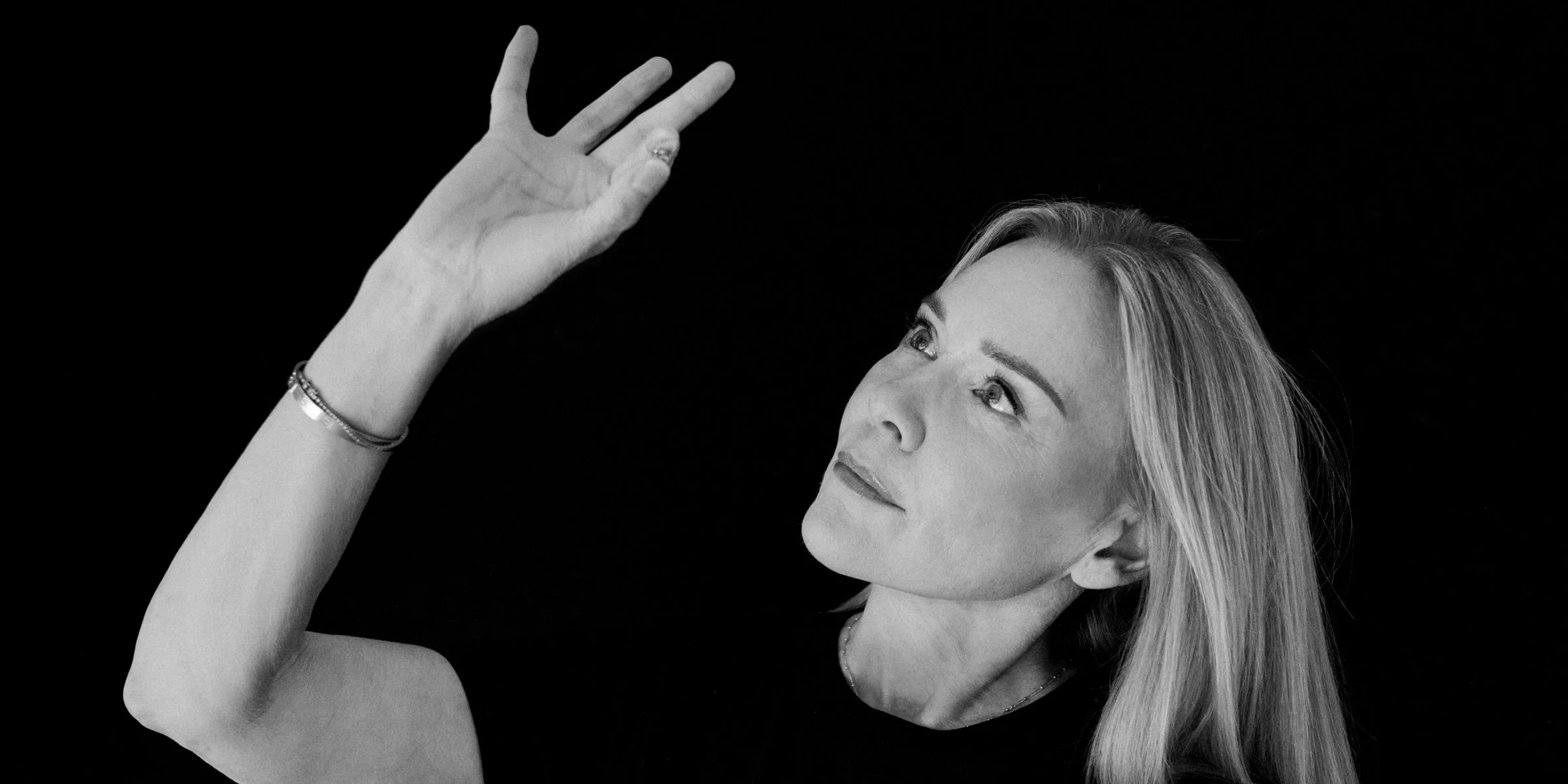En svartvit bild av en kvinna med långt blont hår som tittar uppåt och blickar noggrant på en lins som hon håller mellan fingertopparna.