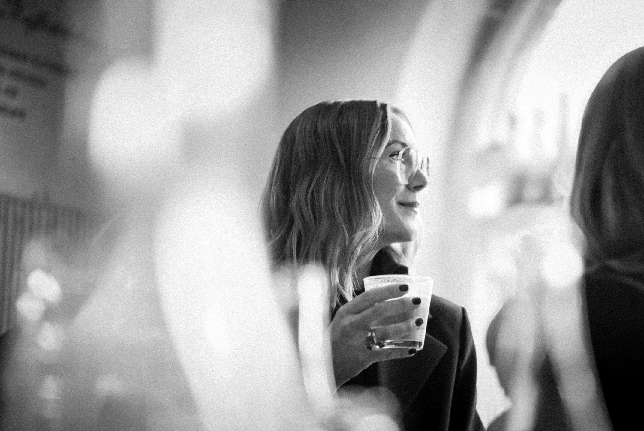 Svartvit bild av två kvinnor på ett kafé, en av dem bär glasögon och håller i ett glas med en cappuccino och tittar ut.