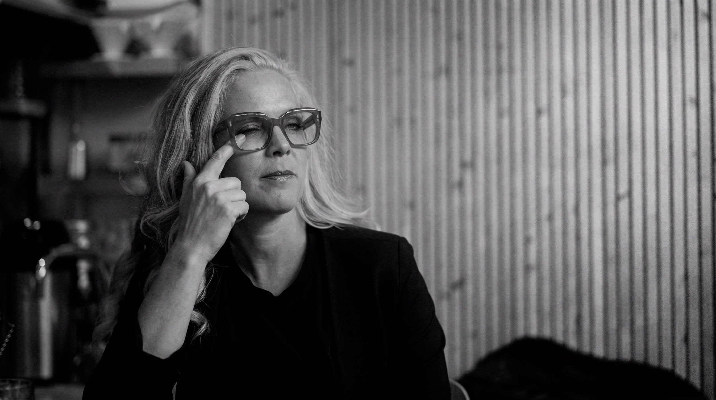 En svartvit bild av en kvinna med blont lockigt hår och stora glasögon, som lite irriterad av torra ögon petar sig i ena ögonvrån.