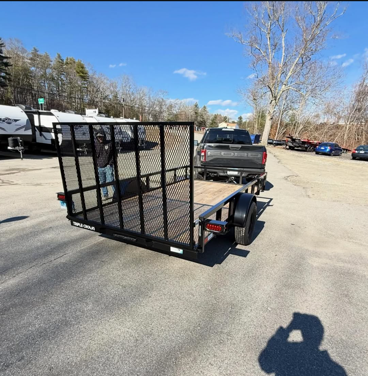 A flatbed trailer attached to a black pickup truck in a parking lot with RVs and other vehicles. The trailer has a black mesh ramp gate and wooden flooring, with one person standing near the back.