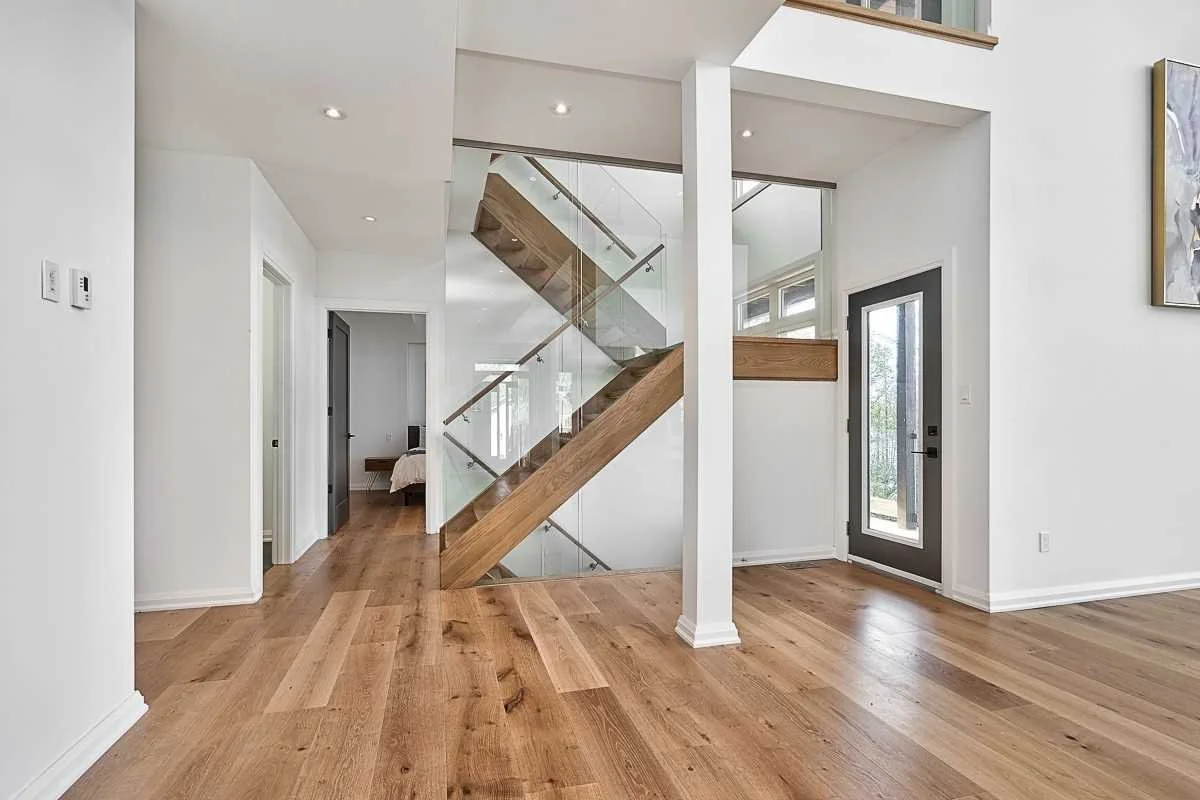 Interior of a modern home featuring a wooden staircase with glass railing, hardwood floors, white walls, a door with glass panel, and a partially visible bedroom.