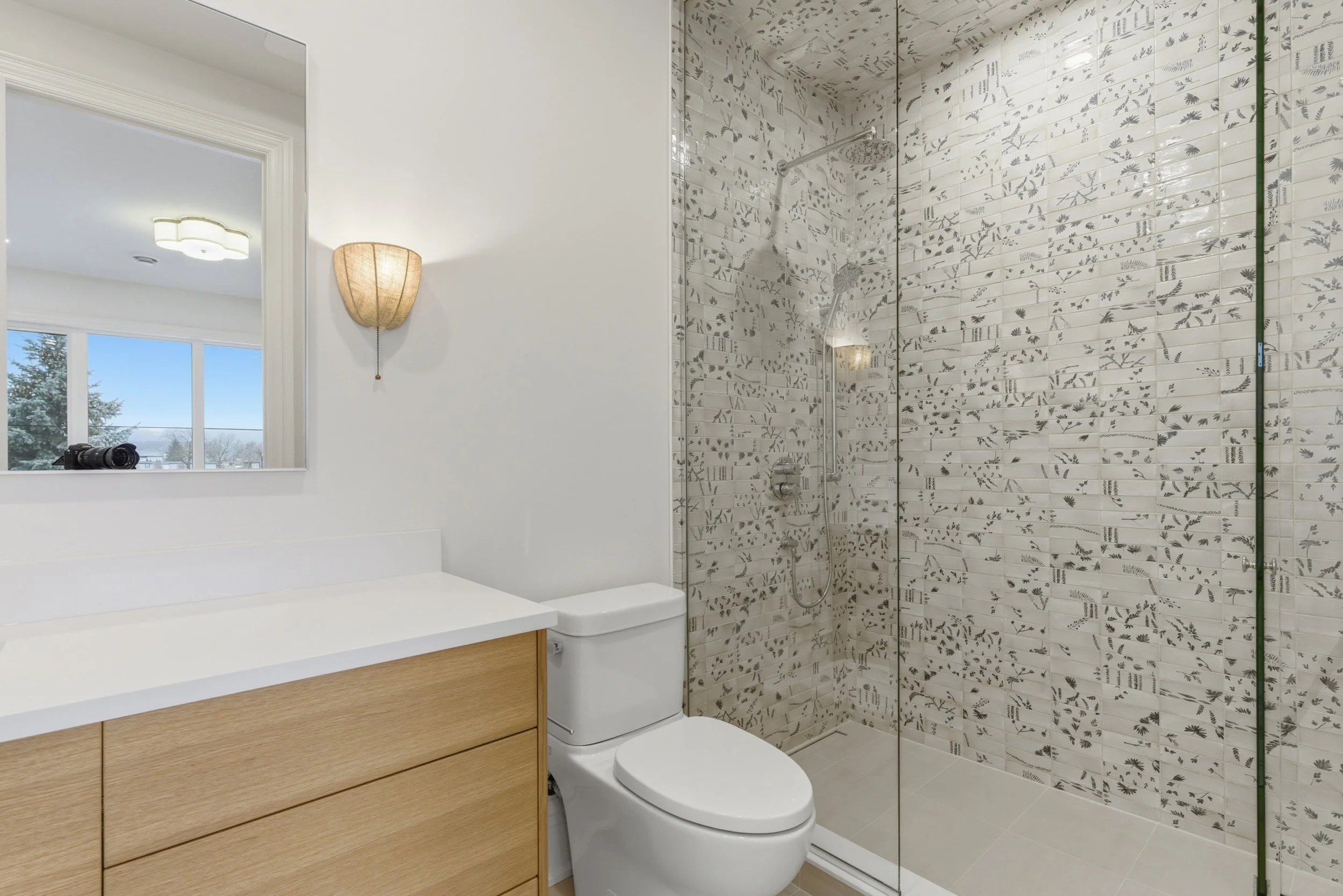 Modern bathroom with wooden vanity, a toilet, and a walk-in shower with patterned tile wall.