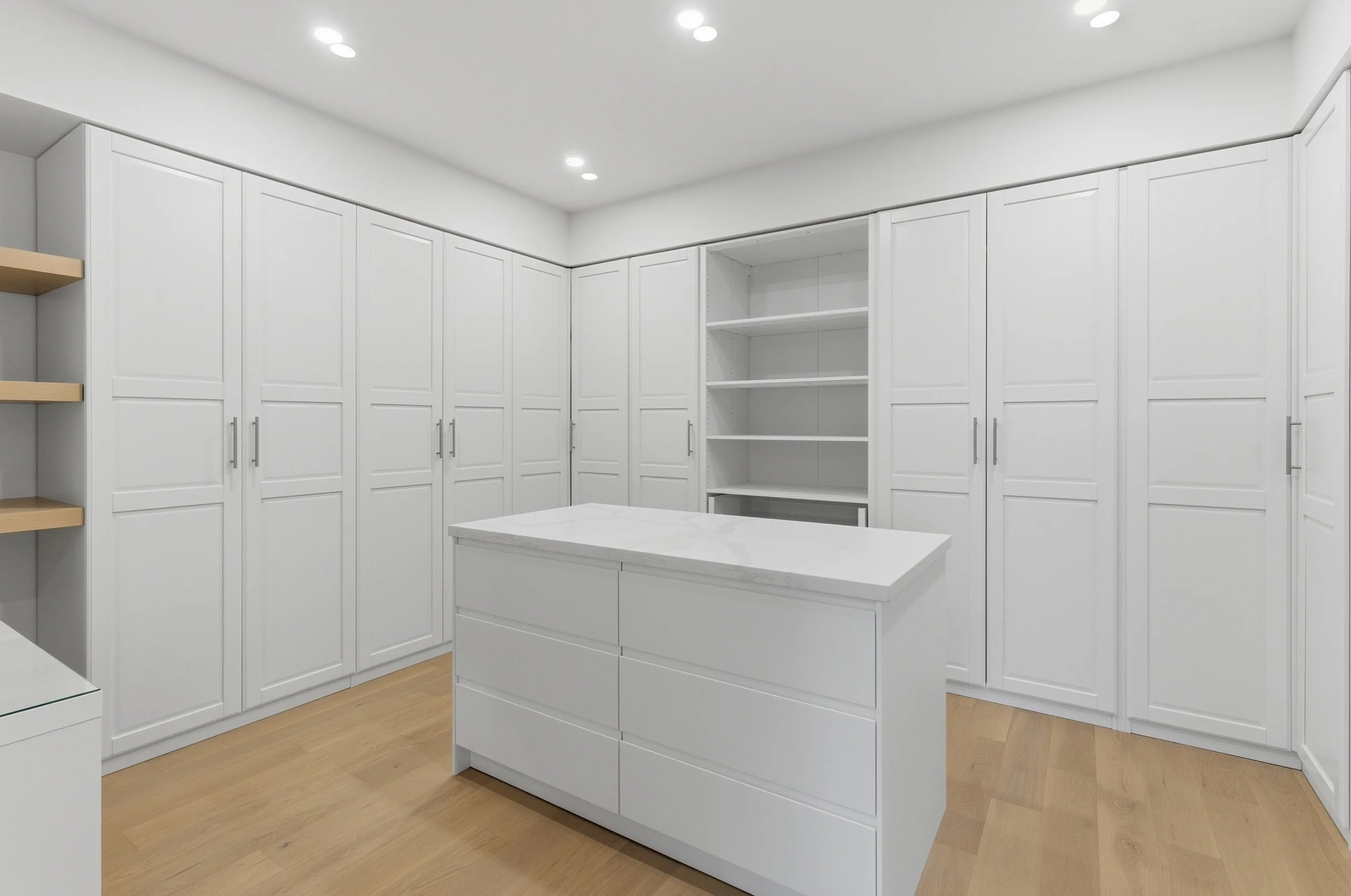 A modern white kitchen with white cabinets, a kitchen island, open shelves, and recessed ceiling lights. Light wood flooring.