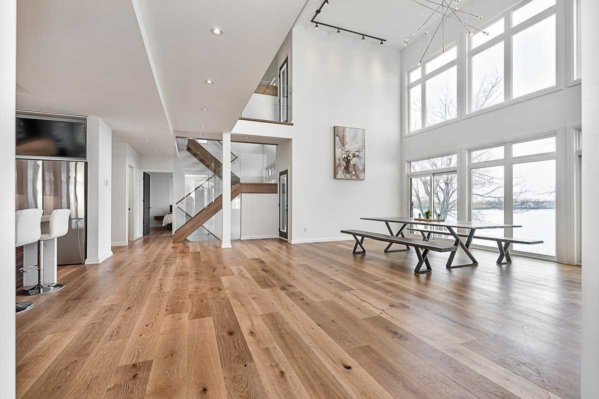 Bright modern living room with large windows, wooden floors, a staircase with wooden steps and glass railing, minimalist wall art, and simple furniture near the windows.