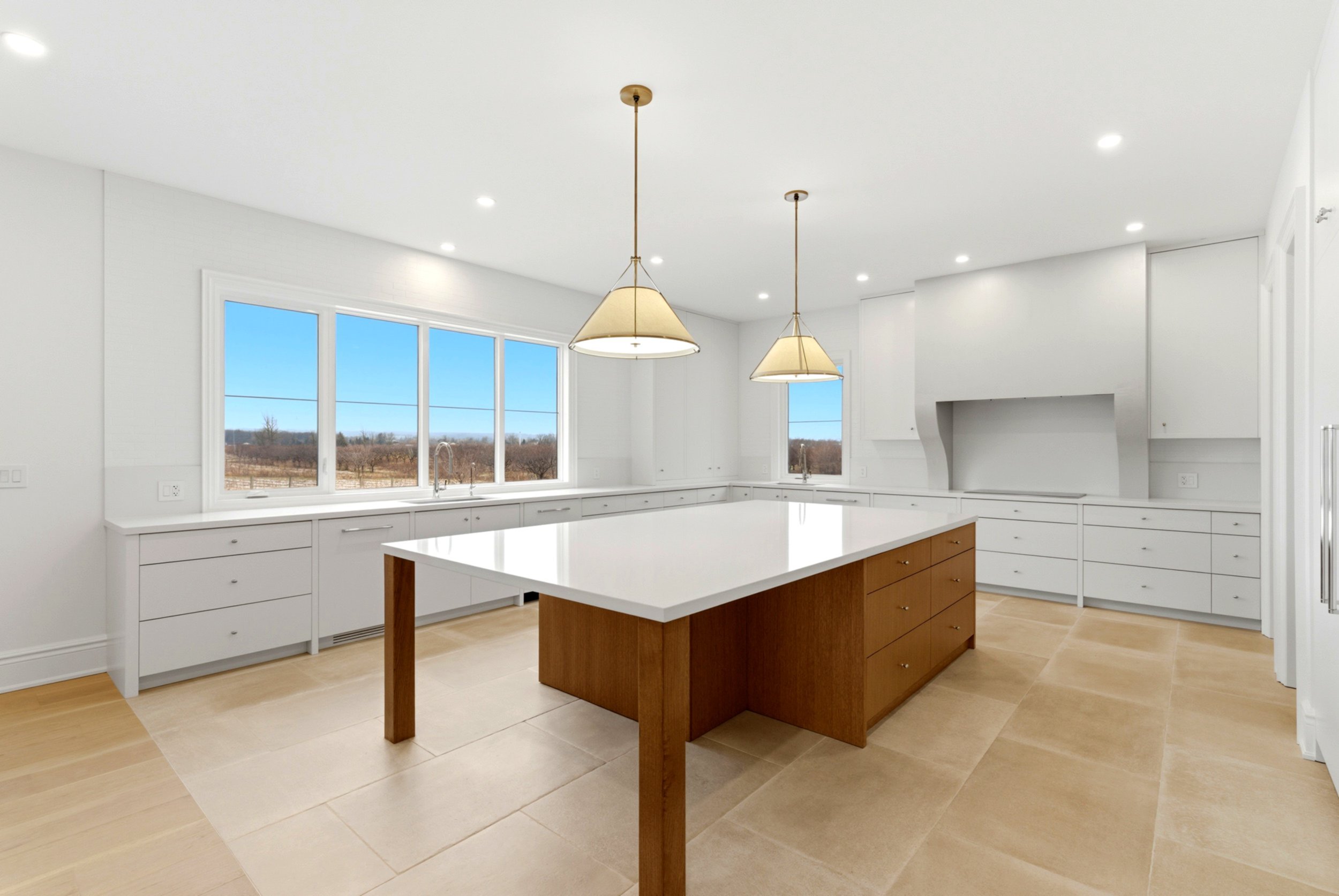 Modern kitchen with white cabinets, a large island with a wooden base and white countertop, large windows showing a rural landscape, and two hanging pendant lights.