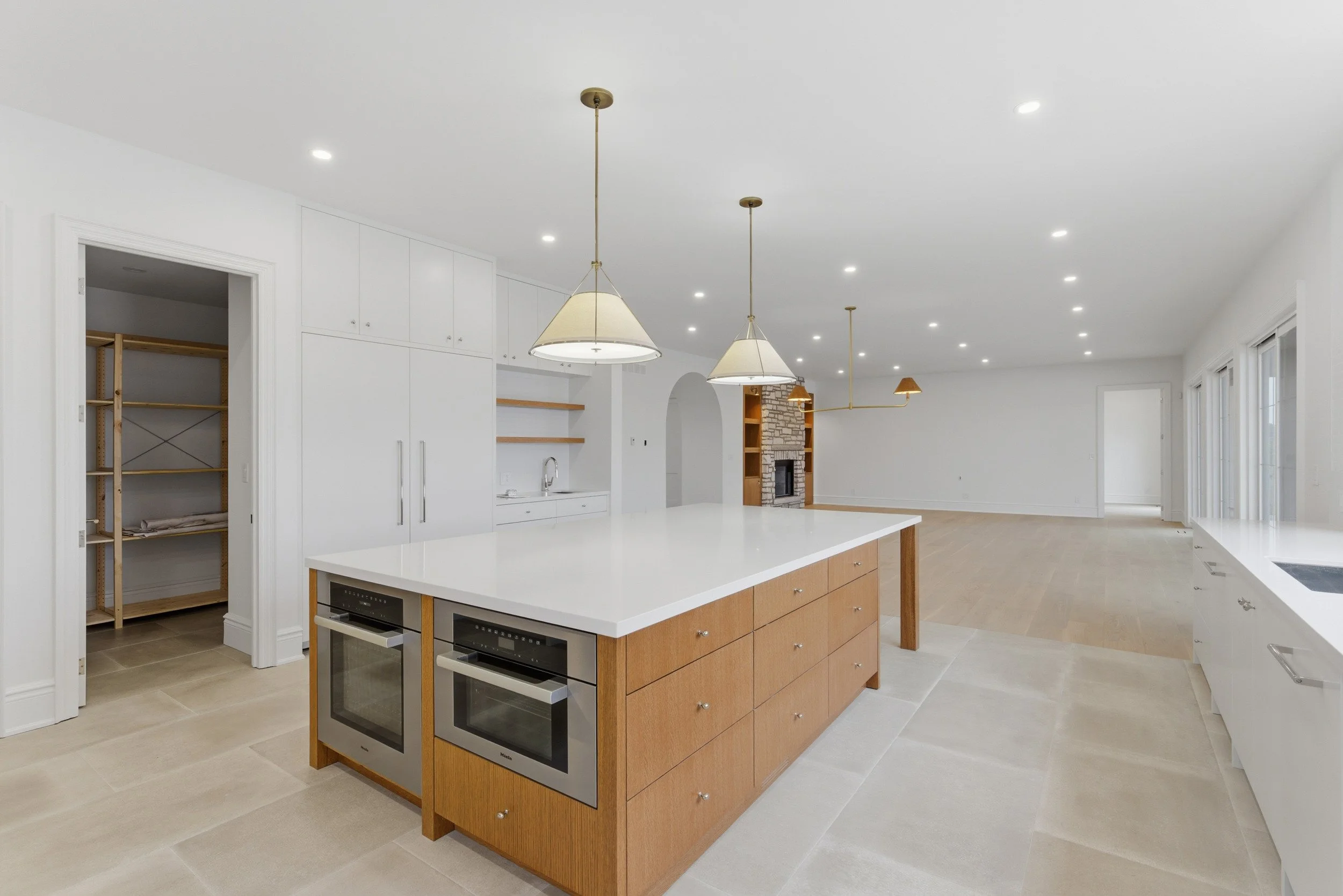Empty modern kitchen with white cabinets, an island with wooden drawers, built-in ovens, and a spacious open living area with a fireplace.