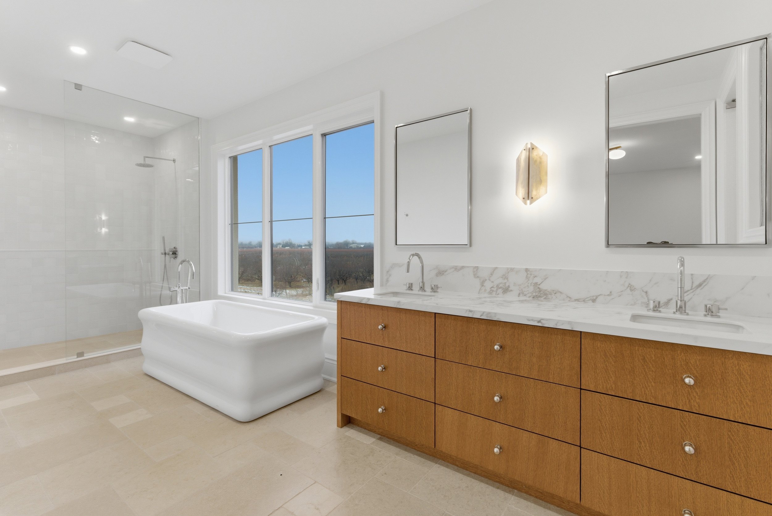 Modern bathroom with a white bathtub, wooden vanity with marble countertop, two mirrors, large window with a view of trees, and a glass-enclosed shower.