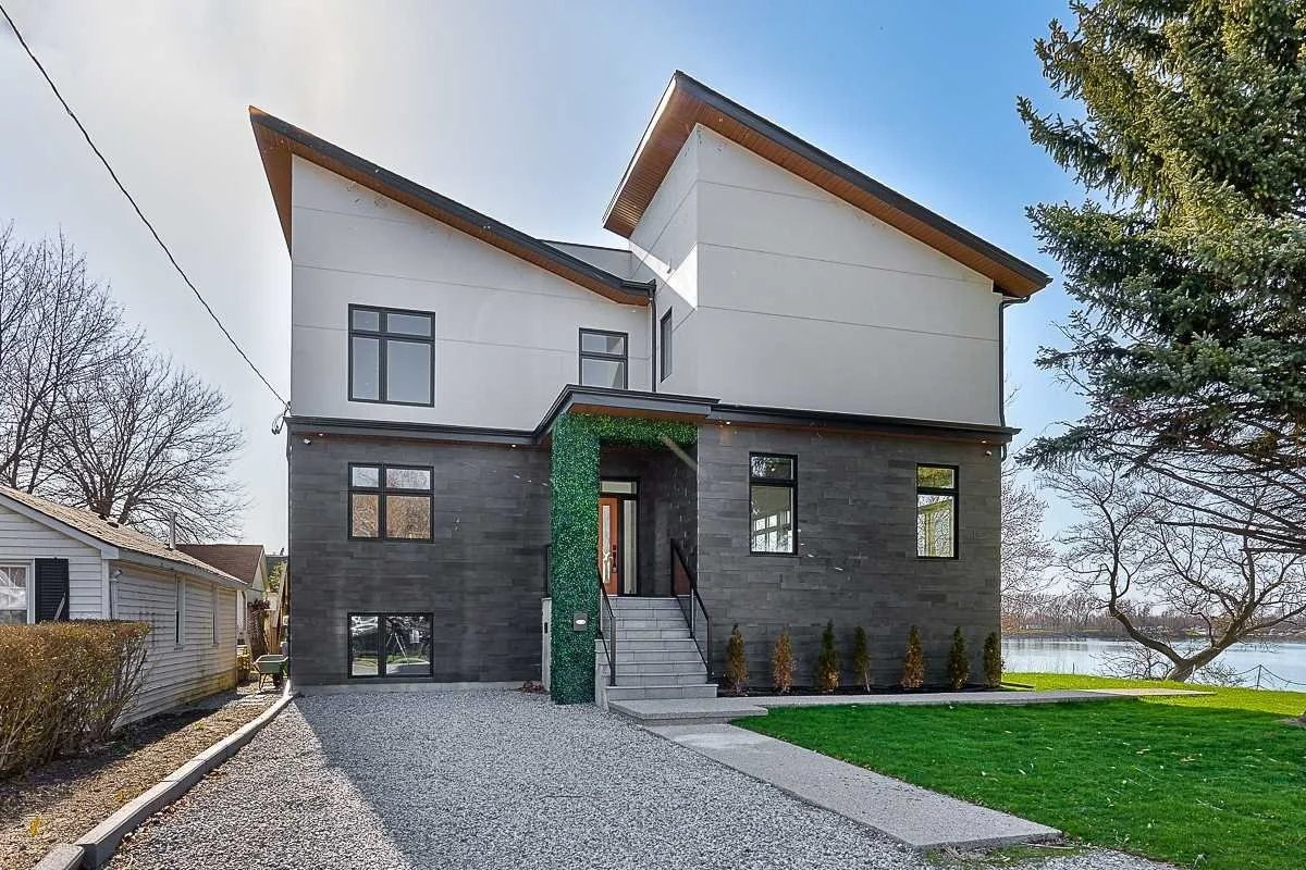 Modern two-story house with dark gray and white exterior, small front porch with stairs, surrounding lawn, gravel driveway, and a pond with trees in the background.