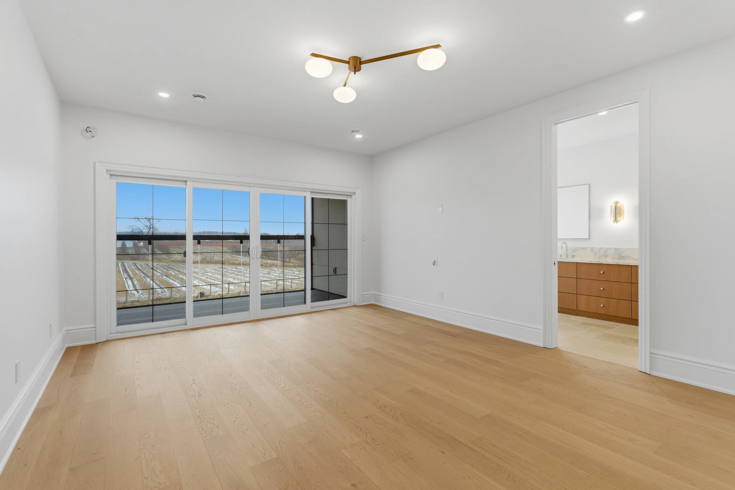 Empty living room with light wood flooring, white walls, large sliding glass door leading to outdoor balcony, and view of a farm field. Connected to bathroom with wooden vanity and marble countertop.