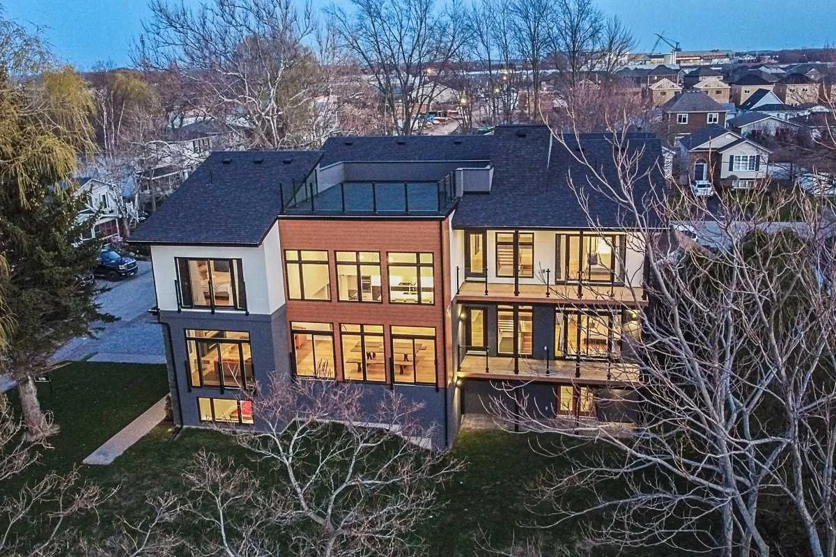 A modern multi-story house with large windows, balconies, and a rooftop deck, situated in a suburban neighborhood with multiple houses and trees.