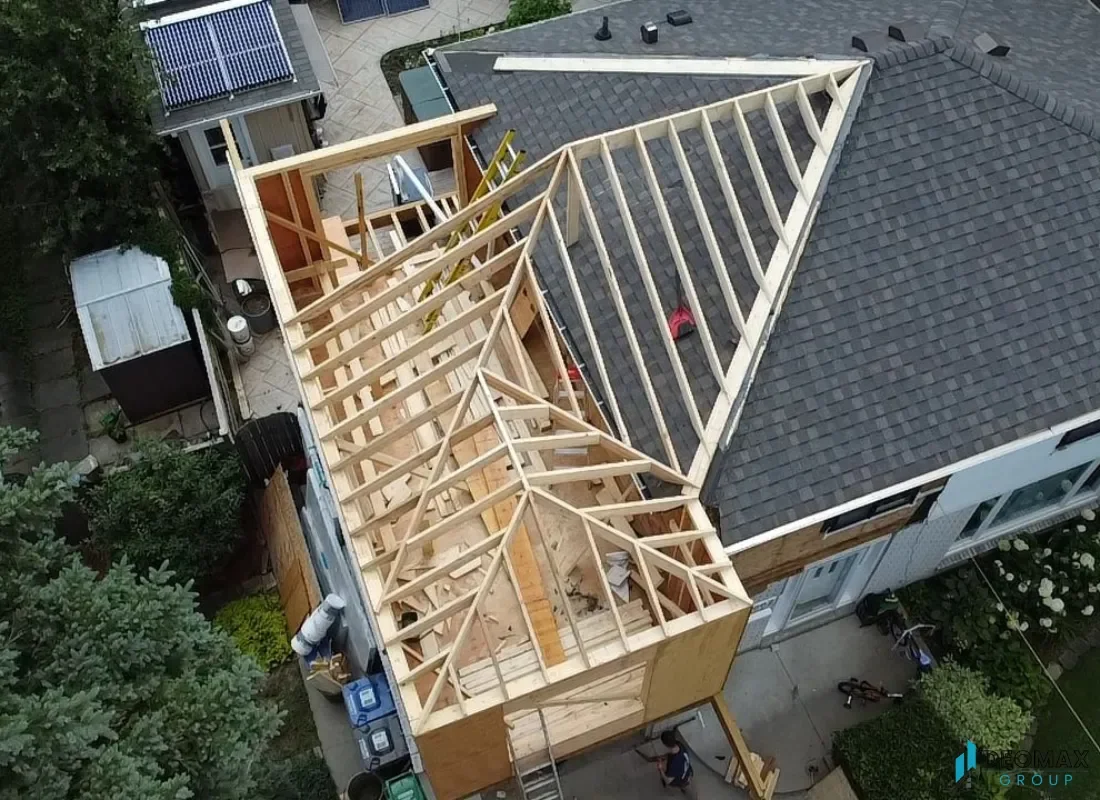 Aerial view of a house under construction with a wood-framed extension being built onto the roof. The existing house has a dark gray shingle roof, and the new extension has a wooden framework. There are construction tools and materials on the ground and on the roof. The property is surrounded by trees, a backyard with a solar panel on a small structure, and a patio area.