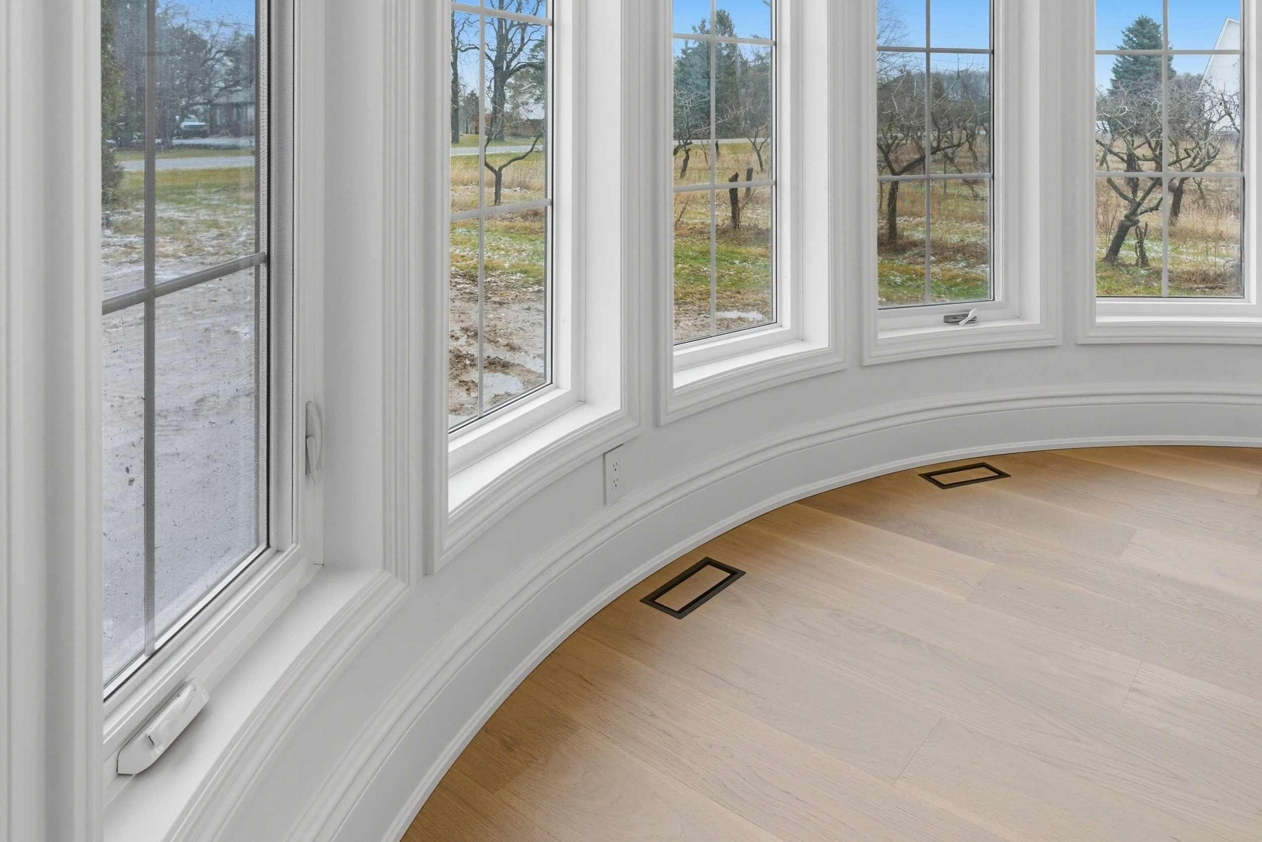 A room with large curved windows, white window frames, and light wood flooring.
