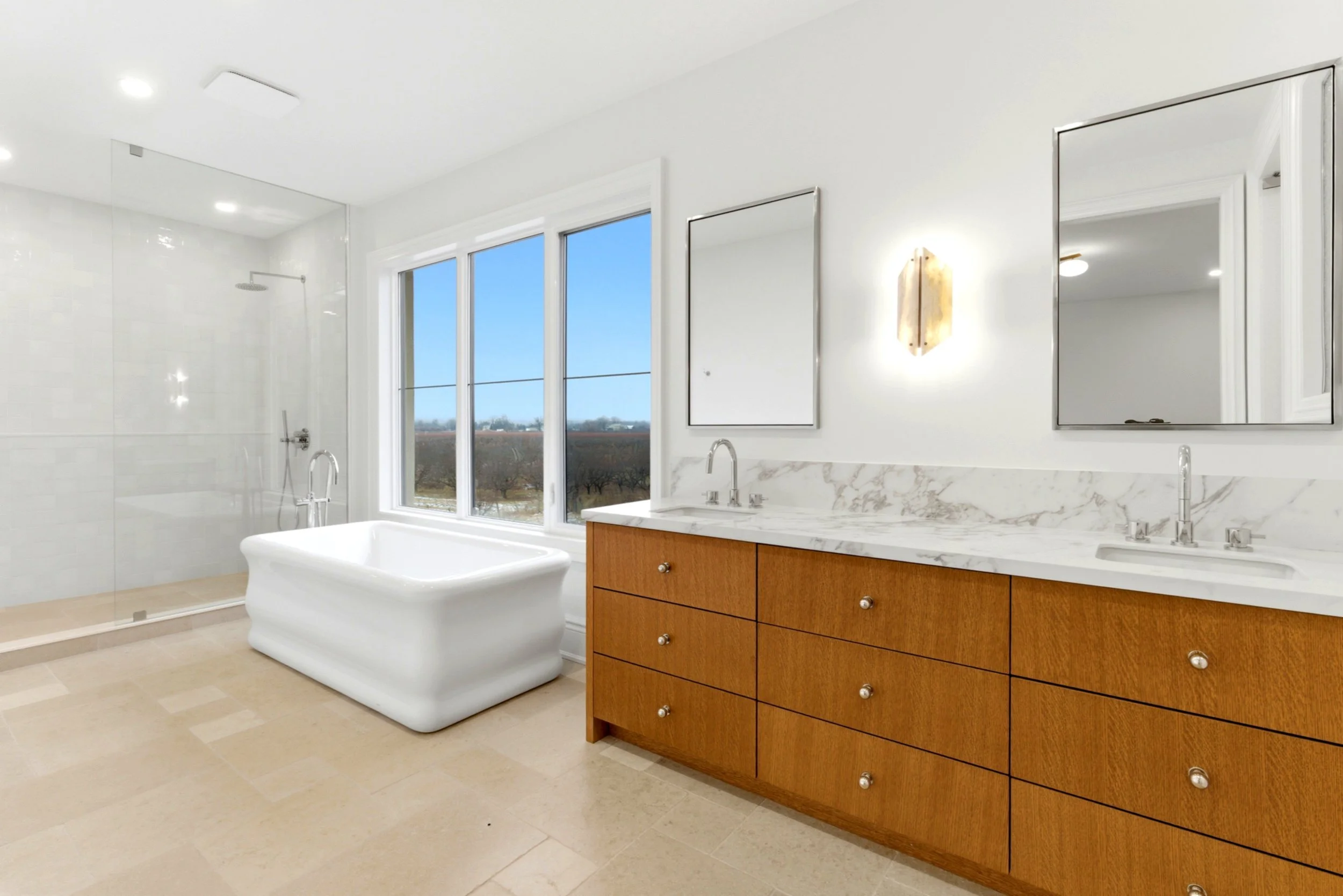 Modern bathroom with walk-in shower, freestanding bathtub, double vanity with marble countertop, two mirrors, and a large window showing a view of trees and blue sky.
