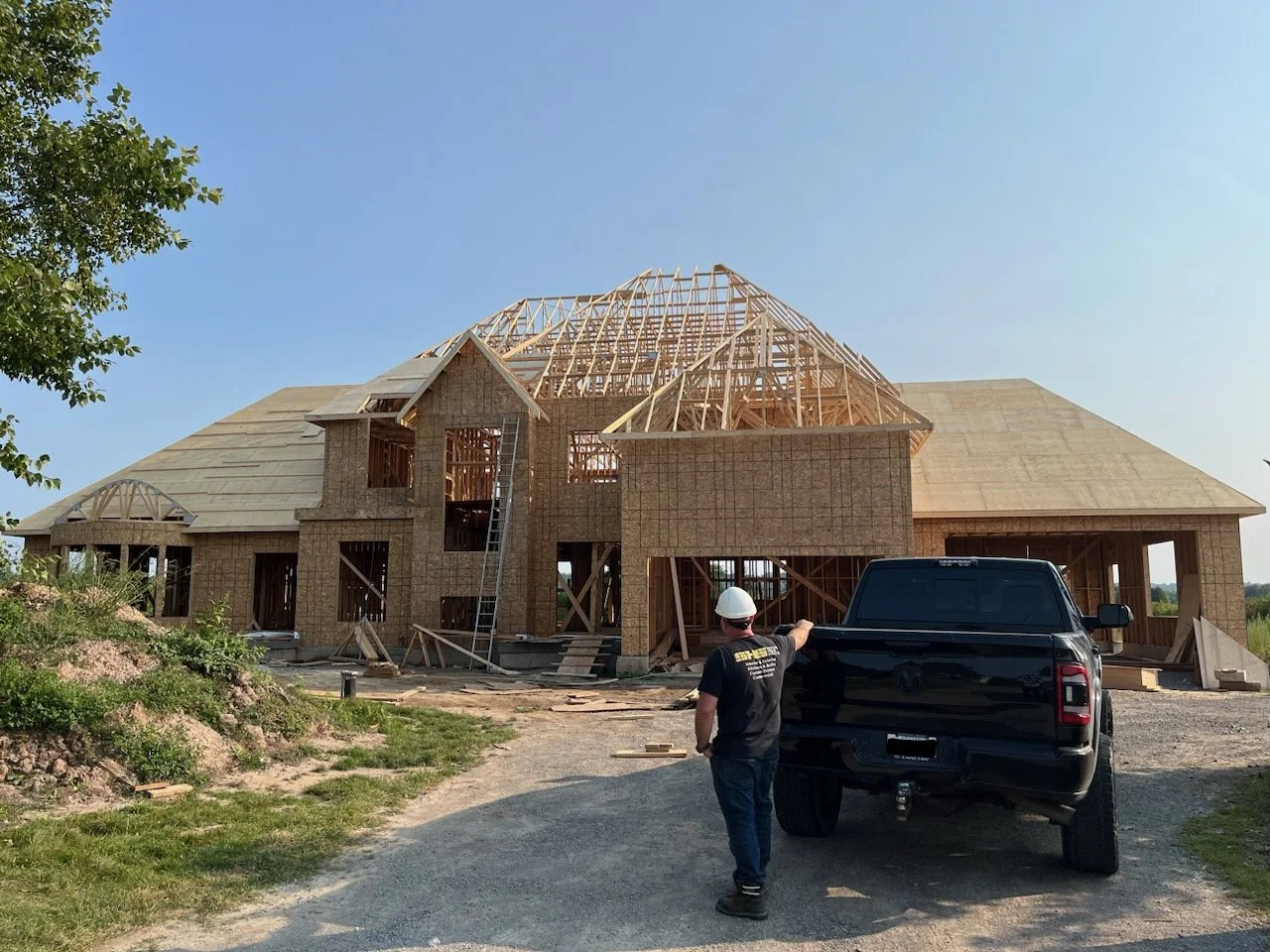 construction site of a large house with wooden framing, a worker in a hard hat pointing at the building, and a black pickup truck in the foreground.