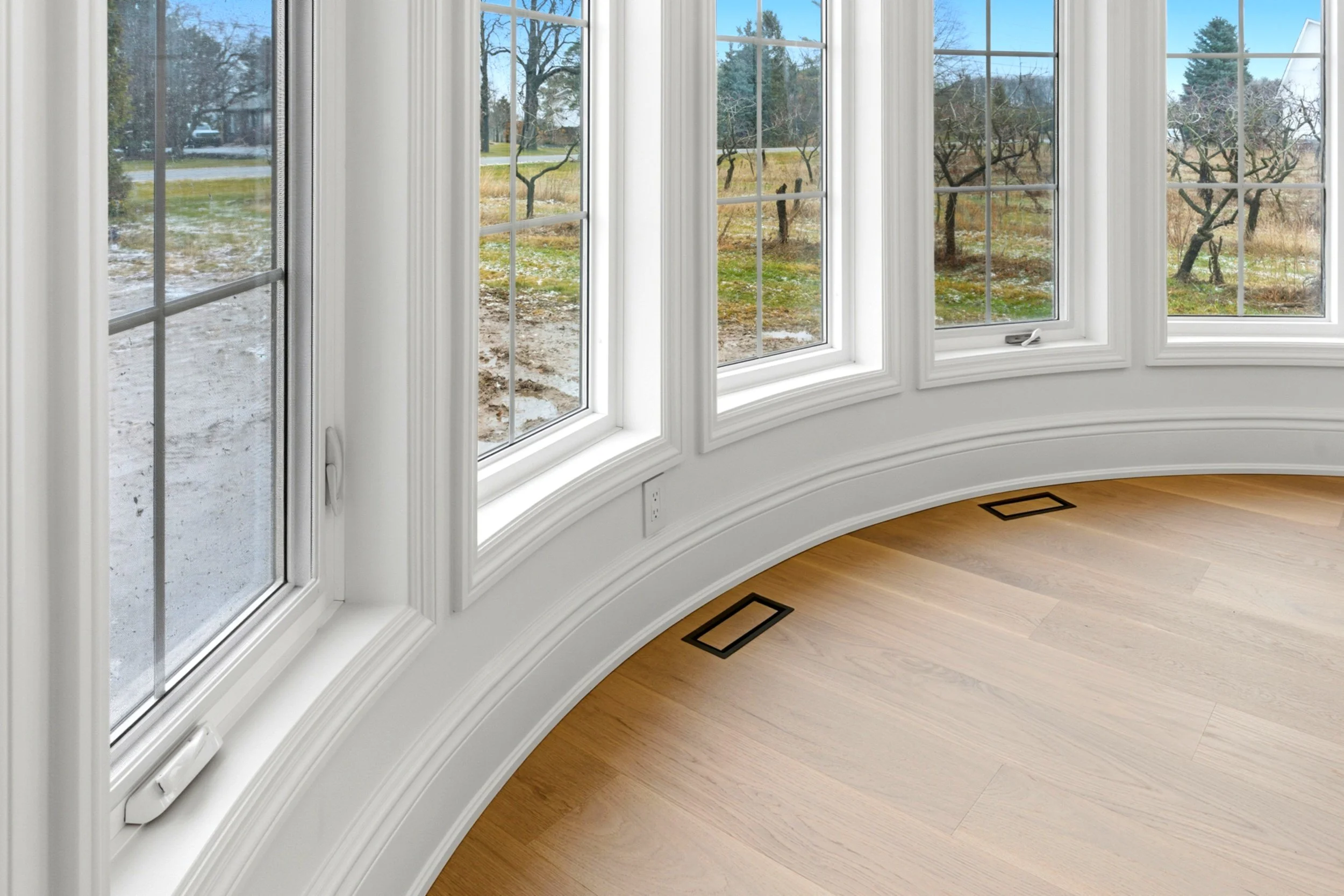 Interior view of a room with a curved wall of large windows revealing a yard with trees and grass outside, wooden flooring, and floor vents.