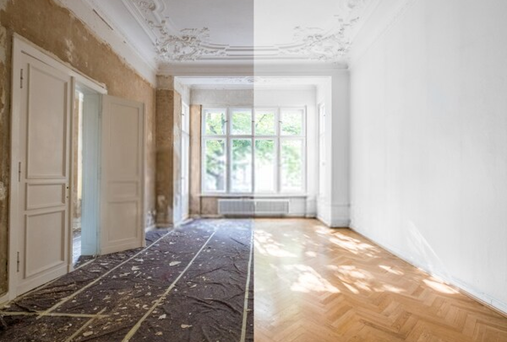 A room showing a renovation contrast: one side with outdated wall and ceiling details, and a damaged floor, the other side with fresh paint and new hardwood flooring, with sunlight streaming through a large window.