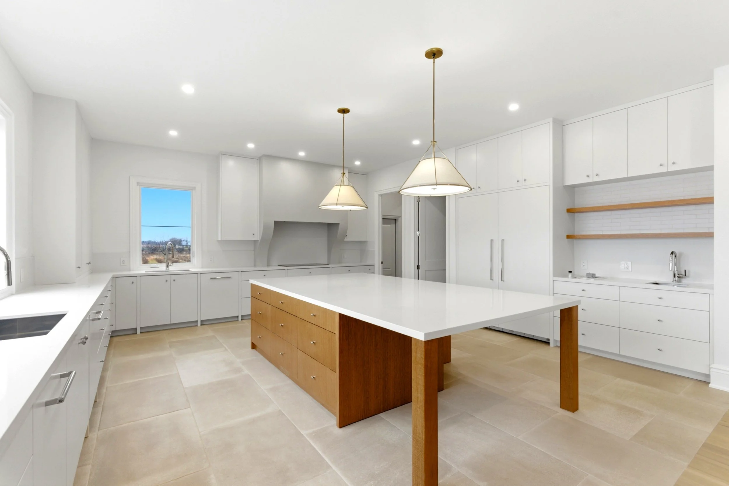 Modern white kitchen with a large island, beige tile flooring, ceiling spotlights, and pendant lights.